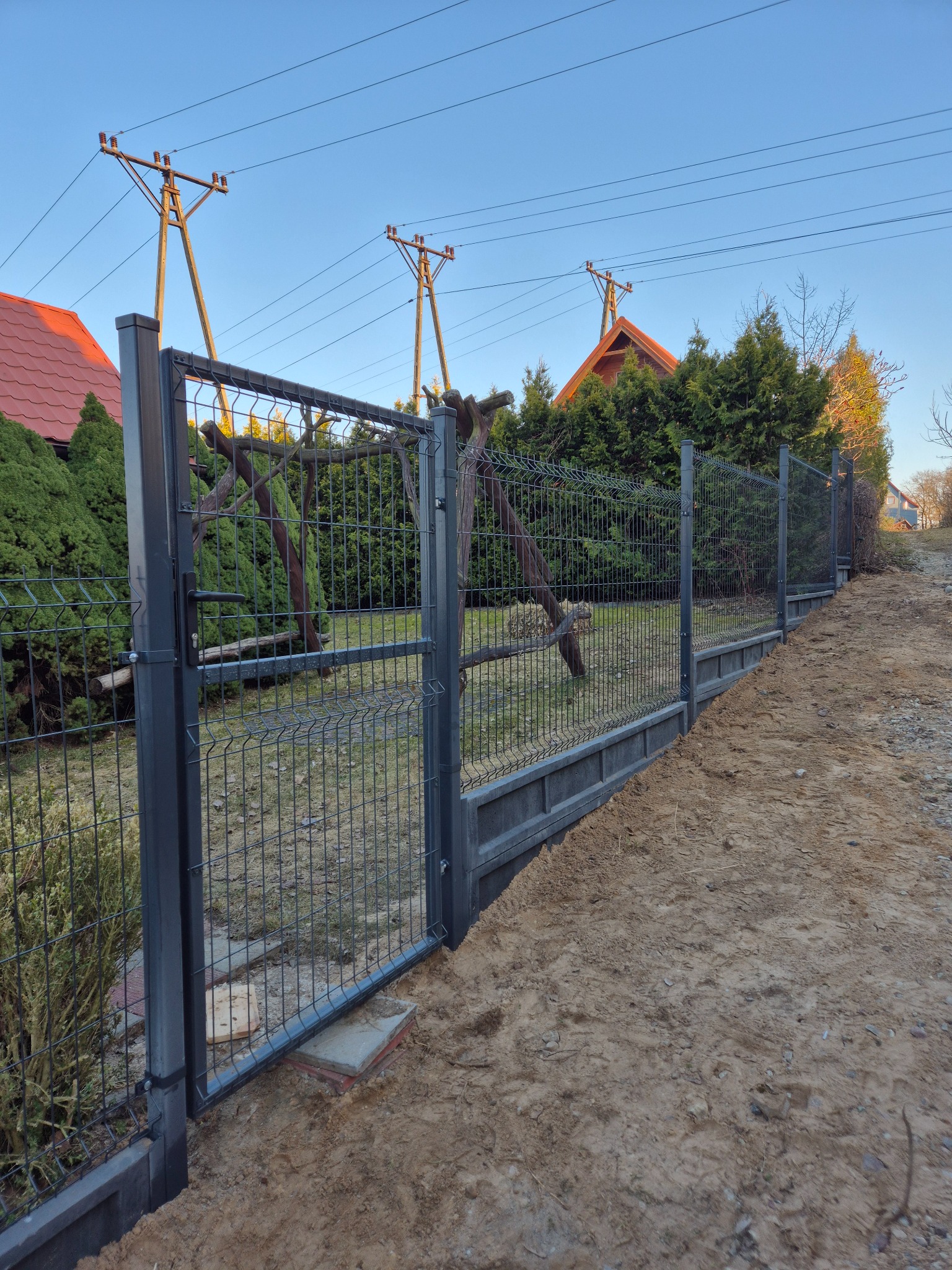 Nowoczesne ogrodzenie panelowe w kolorze grafitowym, z podmurówką betonową, na tle zieleni i błękitnego nieba. Widoczne słupy i przęsła ogrodzenia.