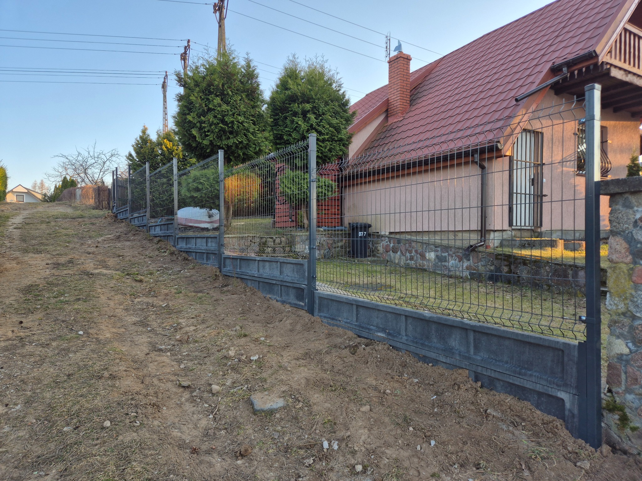 Szary panelowy system ogrodzeniowy z podmurówką betonową, biegnący wzdłuż nierównego terenu działki, z domem w tle. Widok z dołu, wczesne popołudnie.