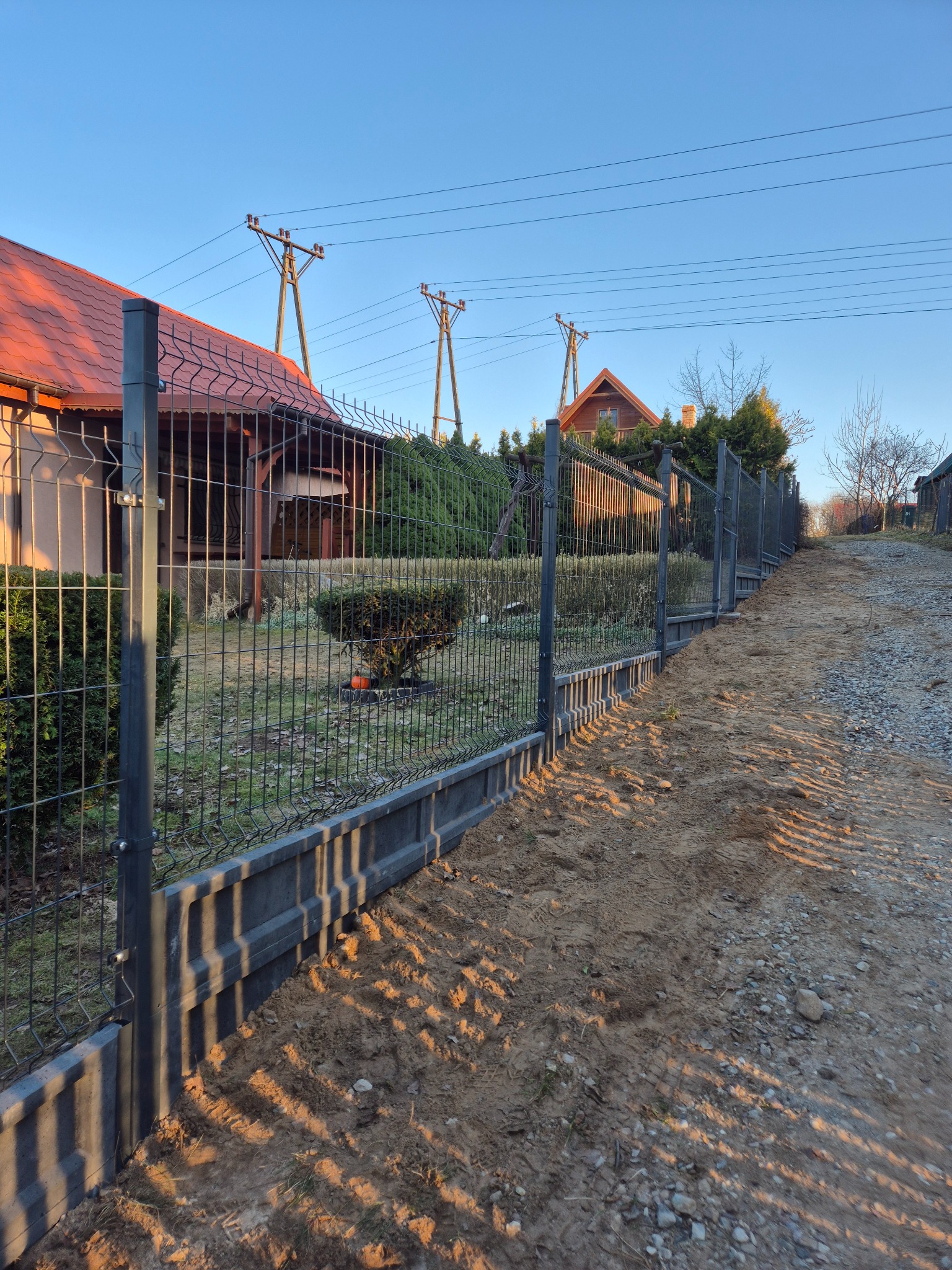 Nowoczesne ogrodzenie panelowe z podmurówką betonową, widok z boku na tle domów i słupów energetycznych w słoneczny dzień. Ciemna siatka i szara podmurówka.