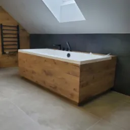 Nowoczesna łazienka na poddaszu z wanną obudowaną drewnopodobnymi panelami, czarną armaturą i grzejnikiem drabinkowym, widoczne okno dachowe.