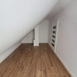 Poddasze z ułożonymi panelami podłogowymi w odcieniach brązu, widoczne białe ściany, skośny sufit, drzwi z przeszkleniami i małe drzwi do schowka.