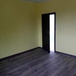 Pusty pok&oacute;j z ż&oacute;łtymi ścianami, ciemną podłogą imitującą drewno i otwartymi ciemnymi drzwiami z srebrną klamką.