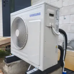 Biała jednostka zewnętrzna klimatyzacji Samsung, zamontowana na betonowych podstawach z gumowymi podkładkami, widoczna na tle ściany budynku z betonu komórkowego i częściowo zieleni