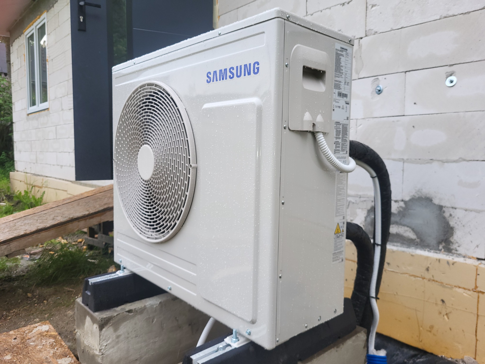 Biała jednostka zewnętrzna klimatyzacji Samsung, zamontowana na betonowych podstawach z gumowymi podkładkami, widoczna na tle ściany budynku z betonu komórkowego i częściowo zieleni