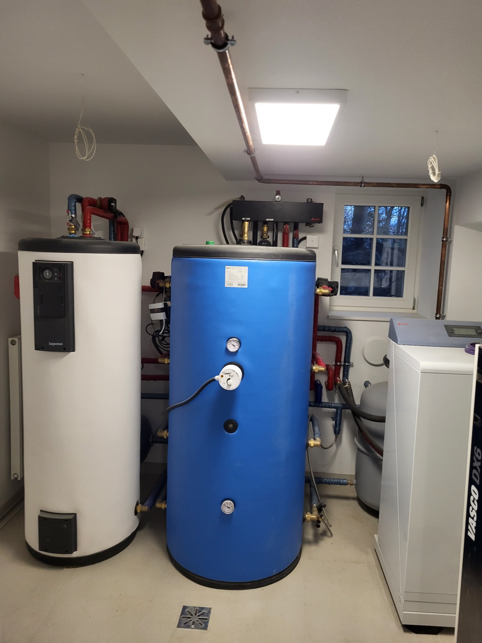Dwa zbiorniki akumulacyjne ciepła, biały i niebieski, z podłączonymi rurami miedzianymi i zaworami, zainstalowane w kotłowni z oknem.
