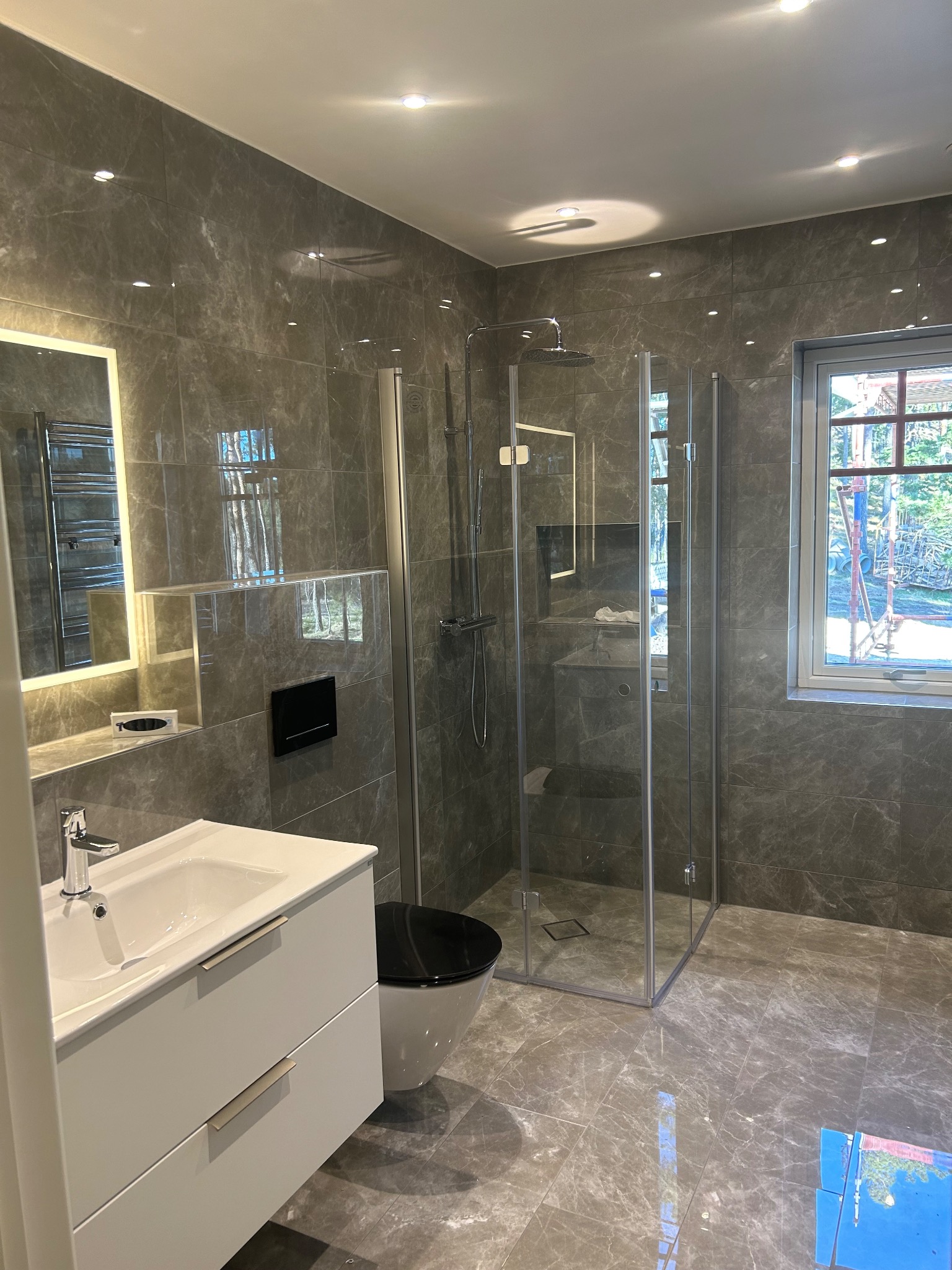 Nowoczesna łazienka wykończona marmurowymi płytkami, z kabiną prysznicową typu walk-in, białą szafką pod umywalkę i podświetlanym lustrem, widok przez okno na zewnątrz.