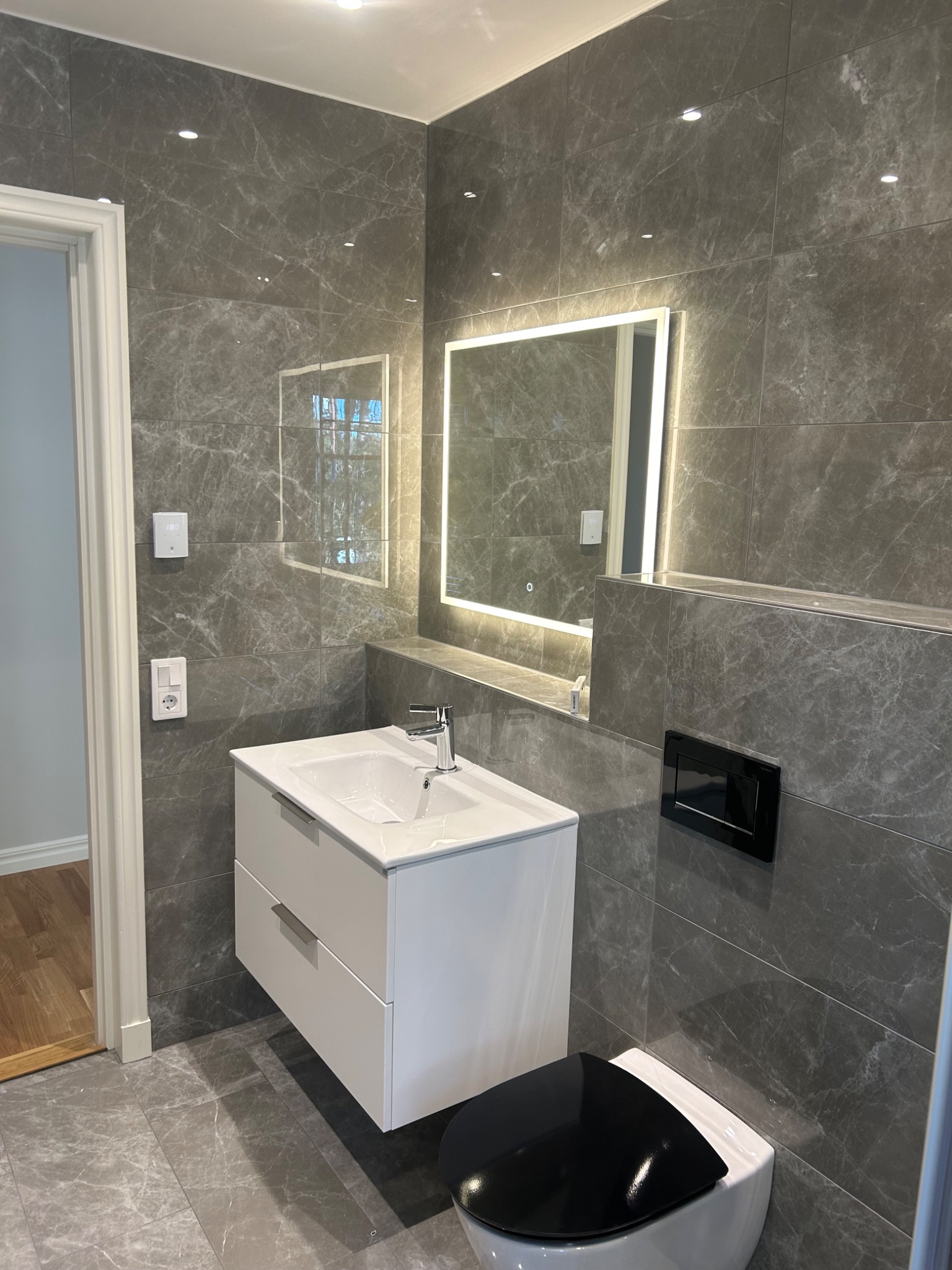 Nowoczesna łazienka wykończona szarymi płytkami imitującymi marmur, z podświetlanym lustrem, białą szafką z umywalką i wiszącą toaletą z czarną deską.