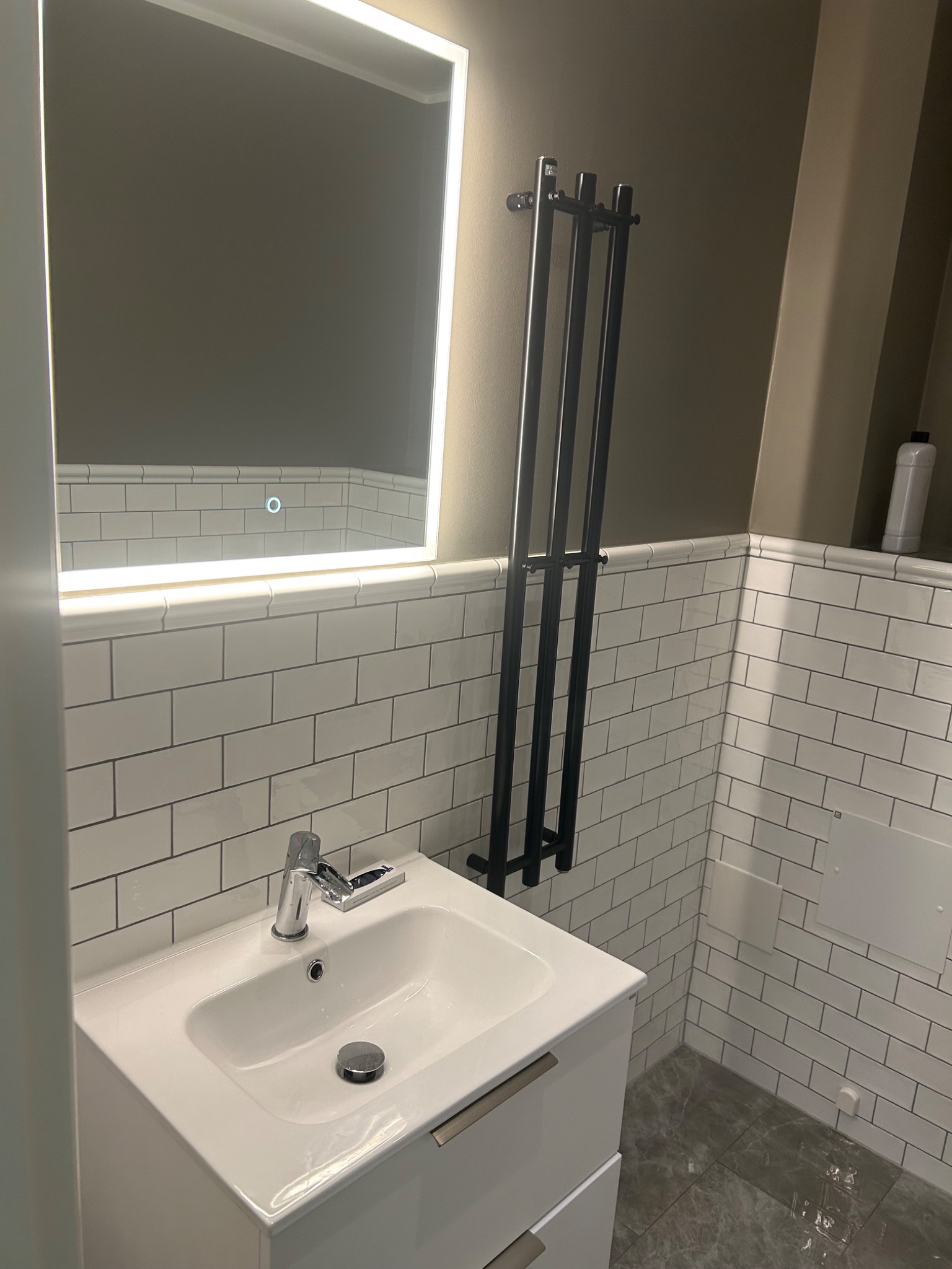 Nowoczesna łazienka z umywalką nablatową na szafce, podświetlanym lustrem z okrągłym włącznikiem dotykowym, grzejnikiem drabinkowym i białymi płytkami imitującymi cegłę.