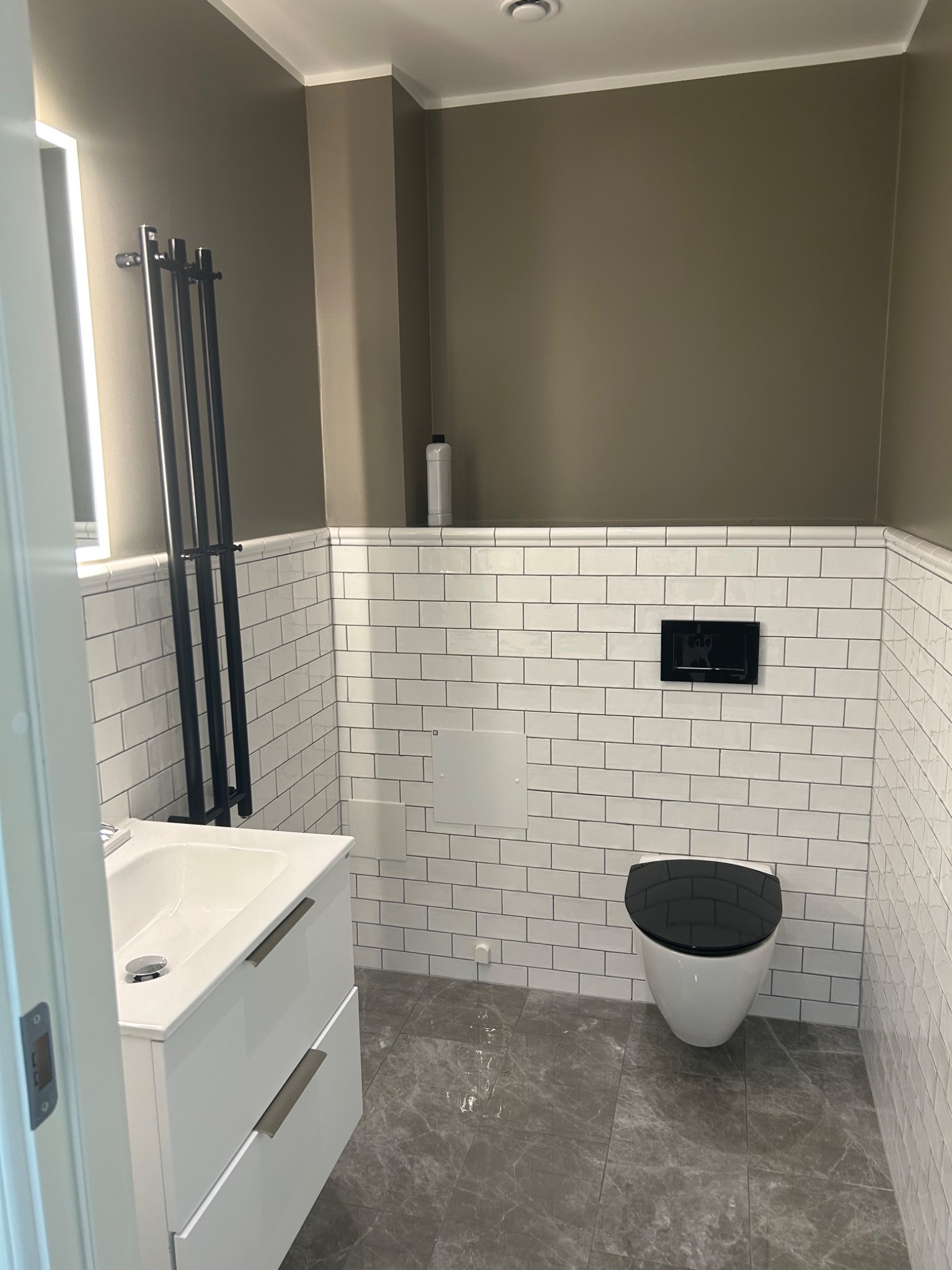 Nowoczesna toaleta z białymi, prostokątnymi płytkami ułożonymi w cegiełkę, szarą podłogą imitującą marmur, wiszącą miską WC z czarną deską, białą szafką z umywalką i czarnym grzejnikiem drabinkowym.