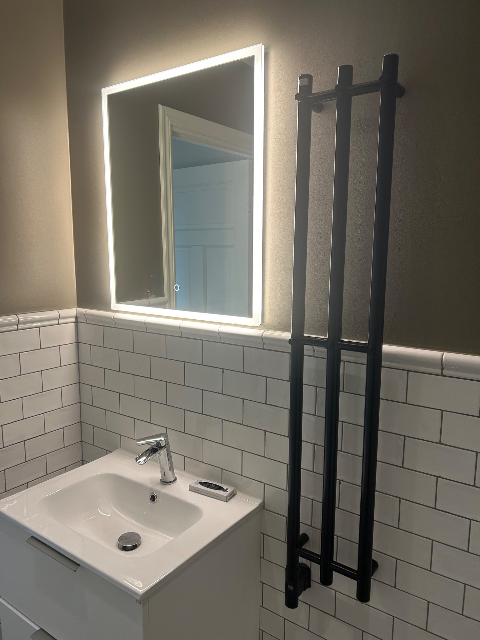 Nowoczesna łazienka z białymi płytkami typu metro, podświetlanym lustrem kwadratowym i czarnym grzejnikiem drabinkowym na ścianie.