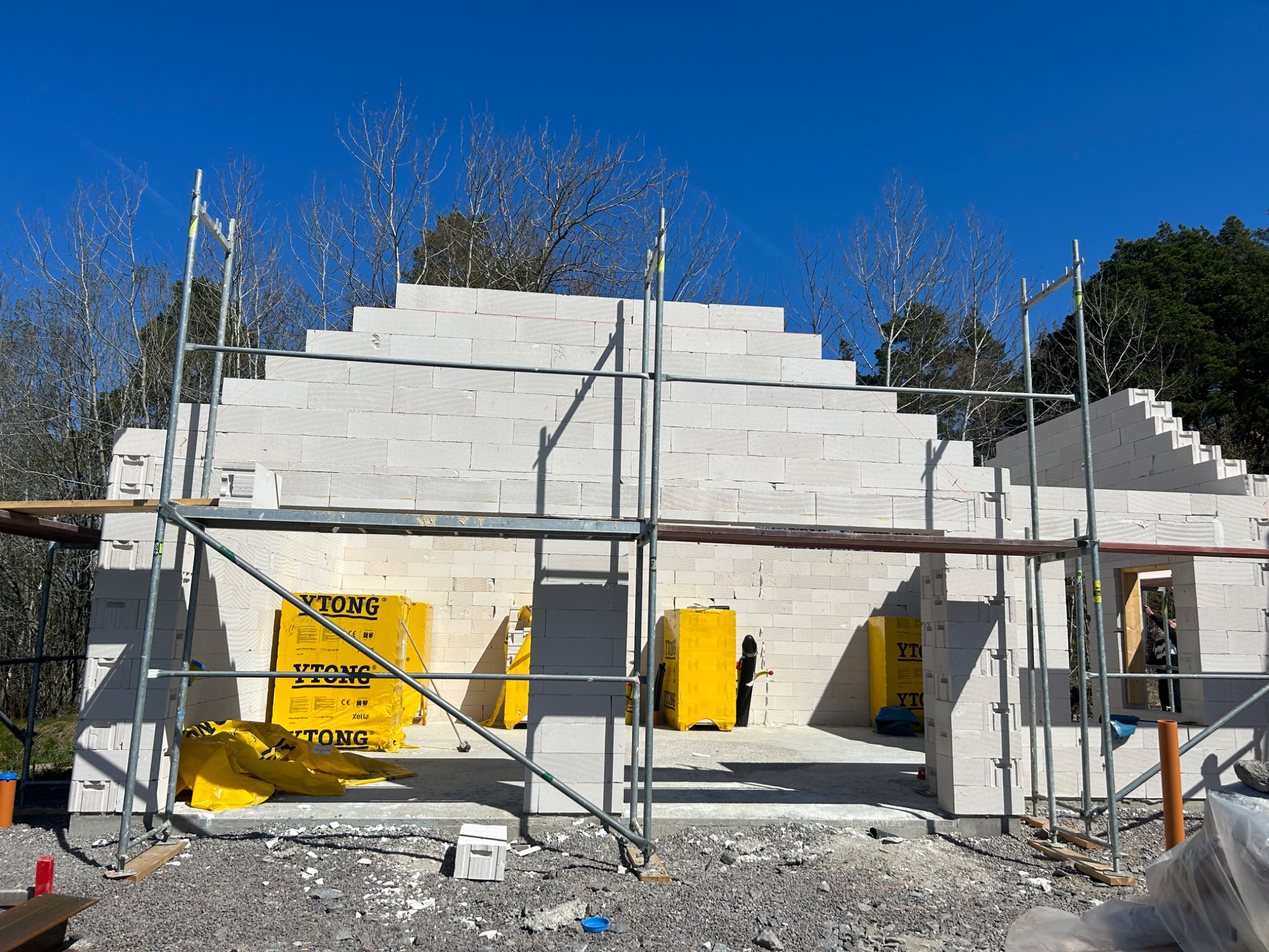 Budowa domu jednorodzinnego z białych bloczków Ytong, widoczne rusztowania i materiały budowlane na placu budowy w słoneczny dzień.