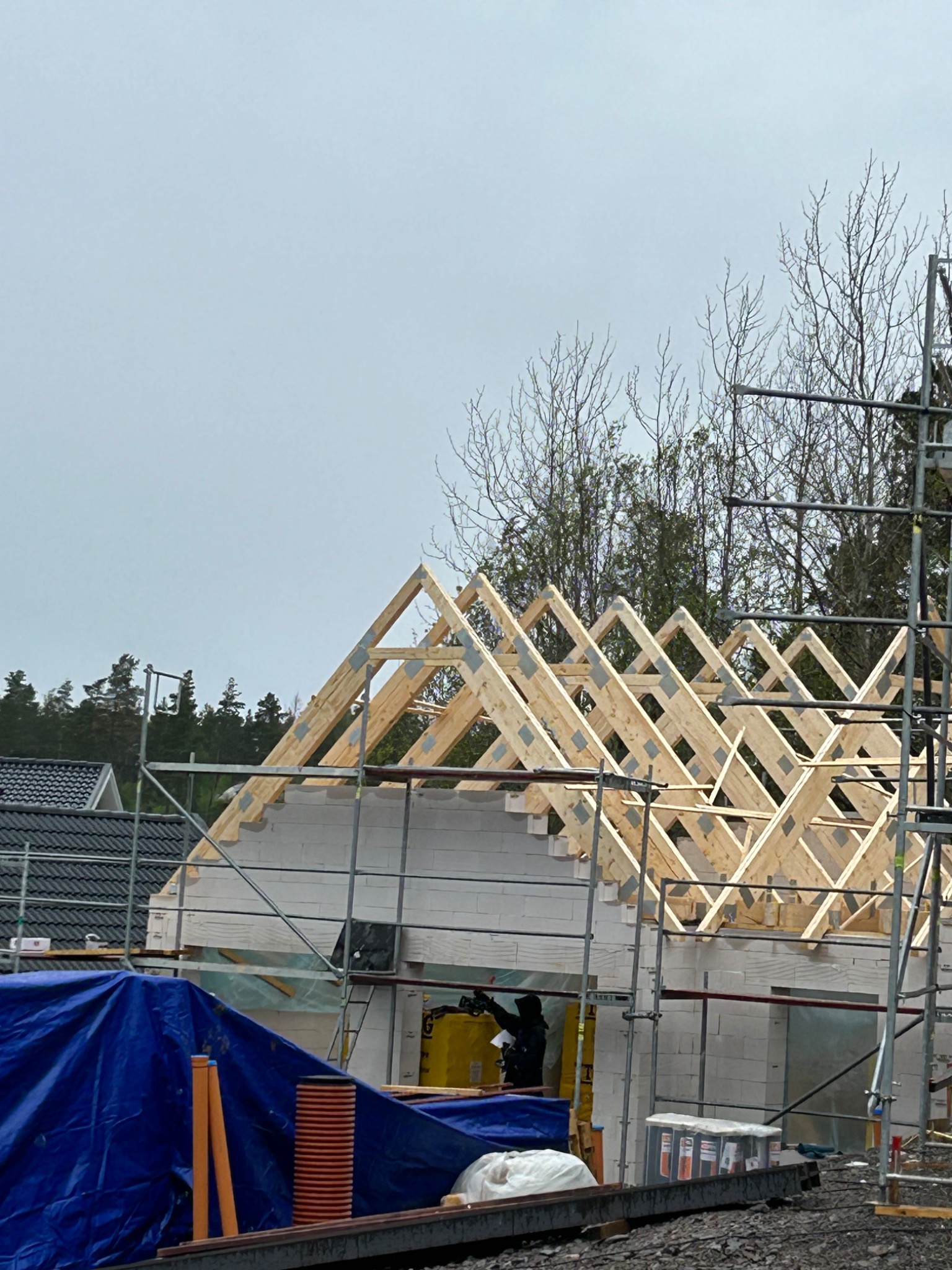 Konstrukcja drewnianego dachu w trakcie montażu na budynku z białych bloczków, widoczne rusztowania i pracownik w ciemnej odzieży, w tle zalesiony teren i pochmurne niebo.