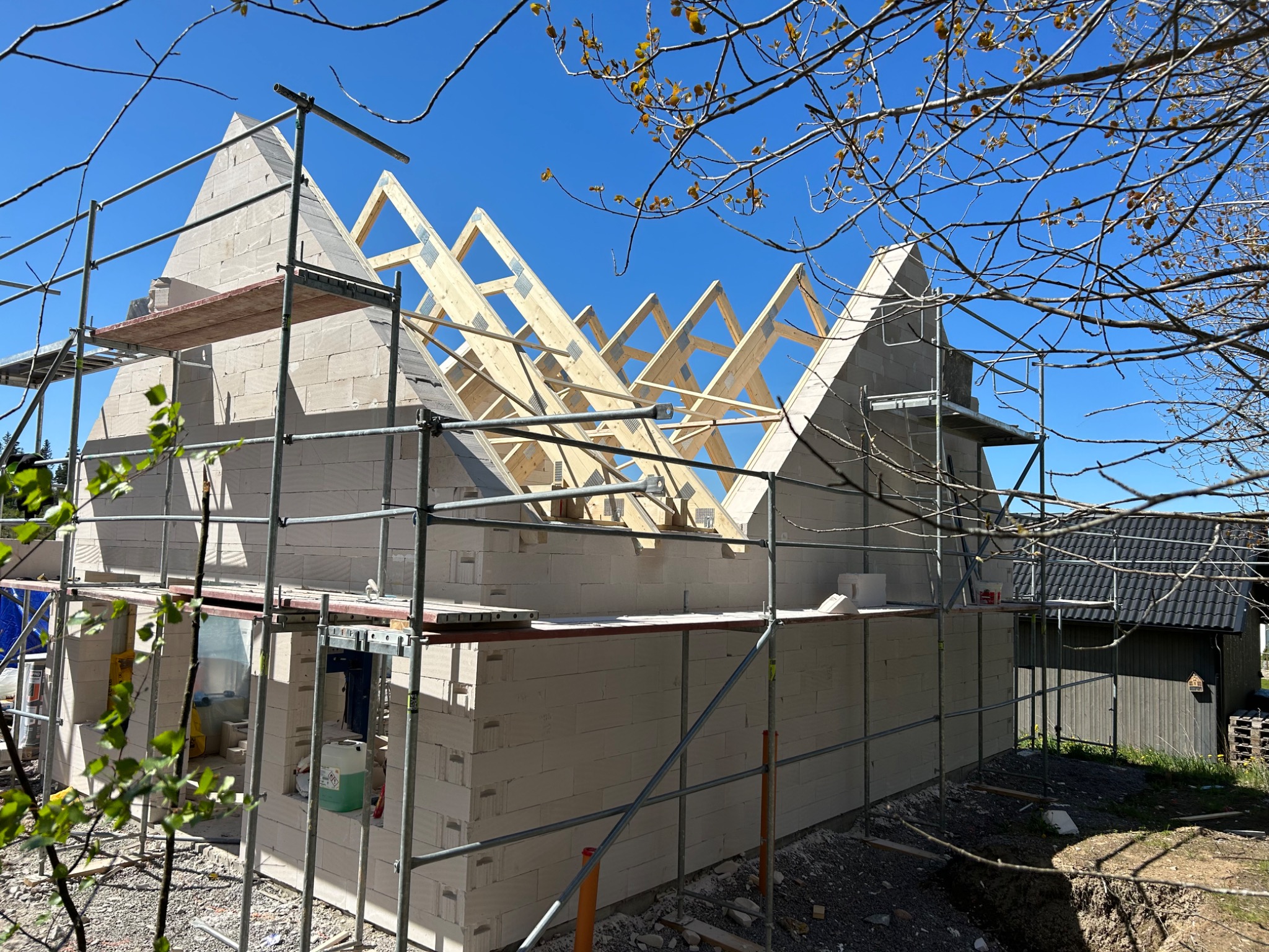 Budowa domu jednorodzinnego z bloczków, widoczne rusztowania, konstrukcja dachu z drewna, błękitne niebo, wiosenne gałęzie drzew