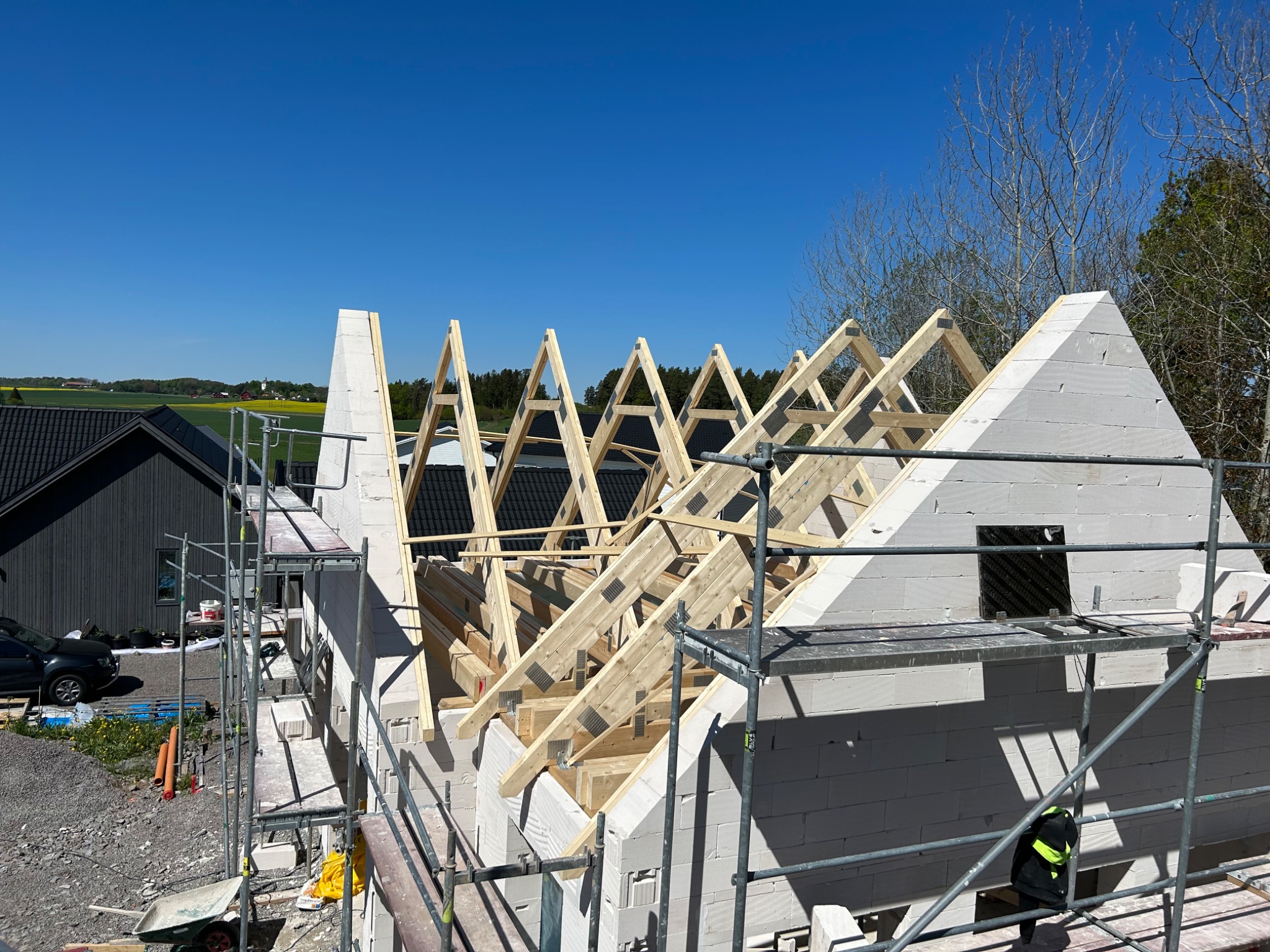 Wznoszenie ścian i więźby dachowej z bloczków betonu komórkowego na rusztowaniu, widok na okoliczne pola i zabudowania w słoneczny dzień.
