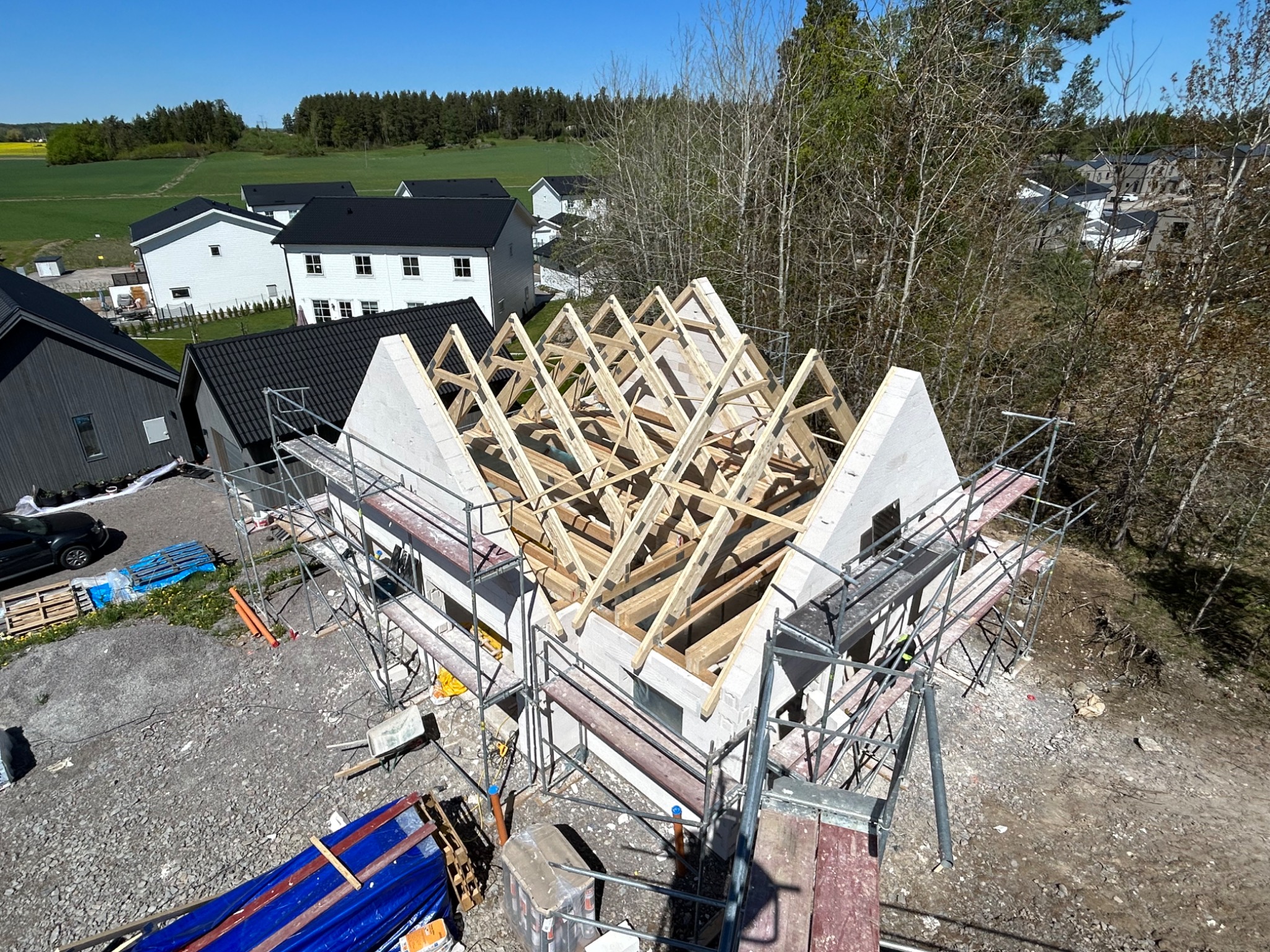 Budowa domu jednorodzinnego: wznoszenie konstrukcji dachu z drewnianych belek na ścianach z białego betonu komórkowego, widoczne rusztowania i otoczenie w postaci innych domów oraz zieleni.