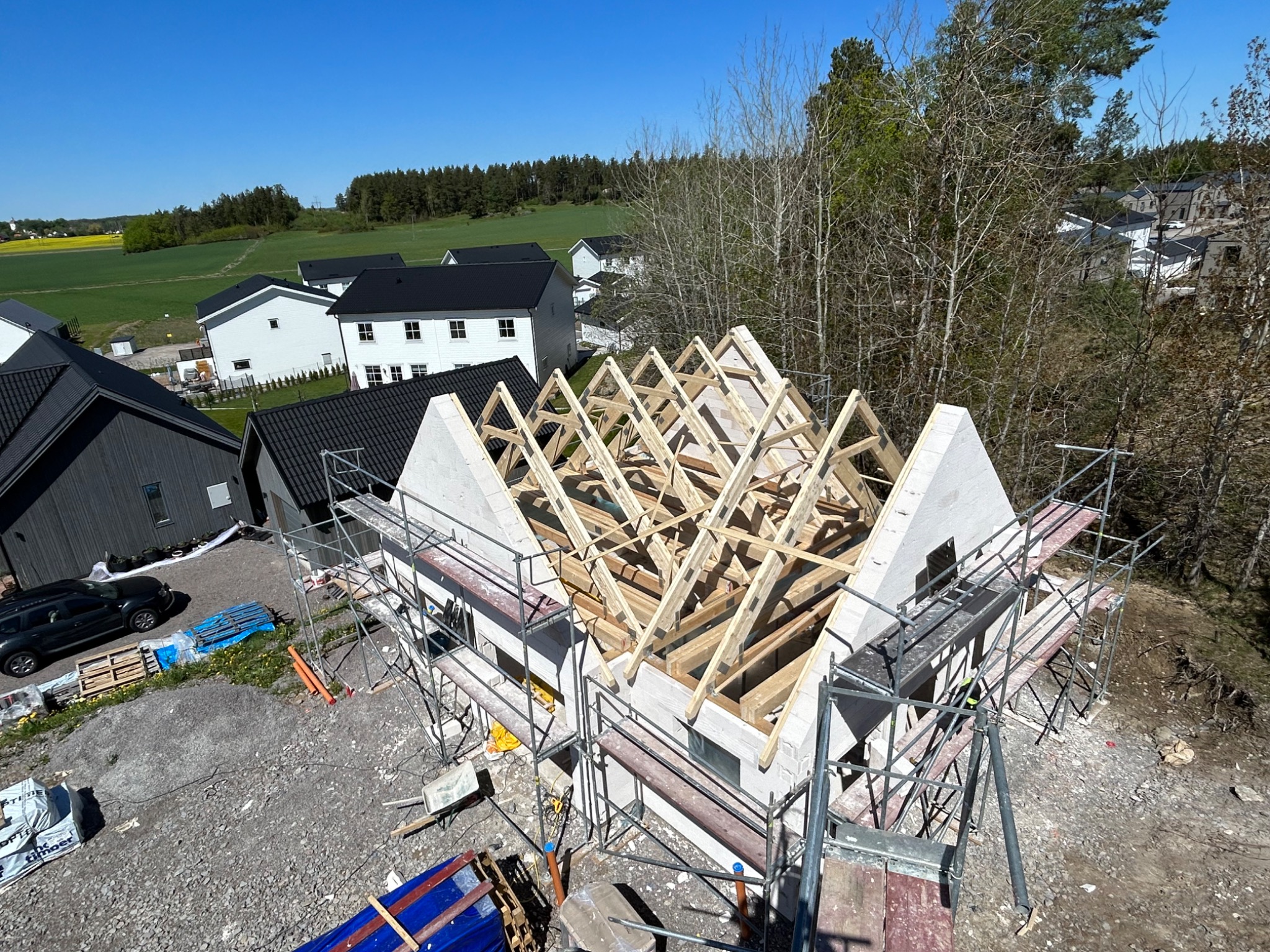 Budowa domu jednorodzinnego z bloczków, widoczne rusztowania, więźba dachowa w trakcie montażu, w tle zabudowa jednorodzinna i pola uprawne.