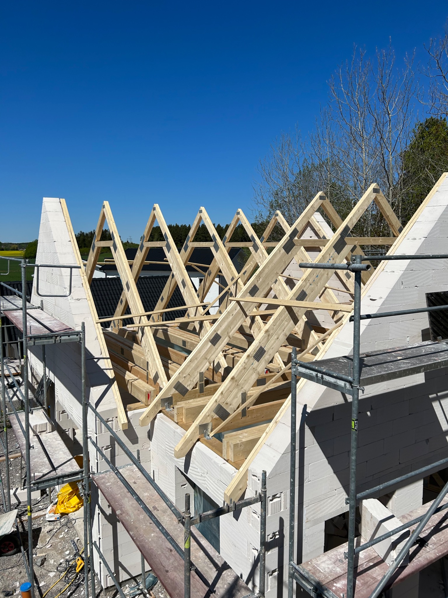 Budowa domu jednorodzinnego: wznoszenie konstrukcji dachu z drewnianych krokwi i ścian z białych bloczków, widoczne rusztowania i narzędzia na placu budowy, słoneczny dzień.