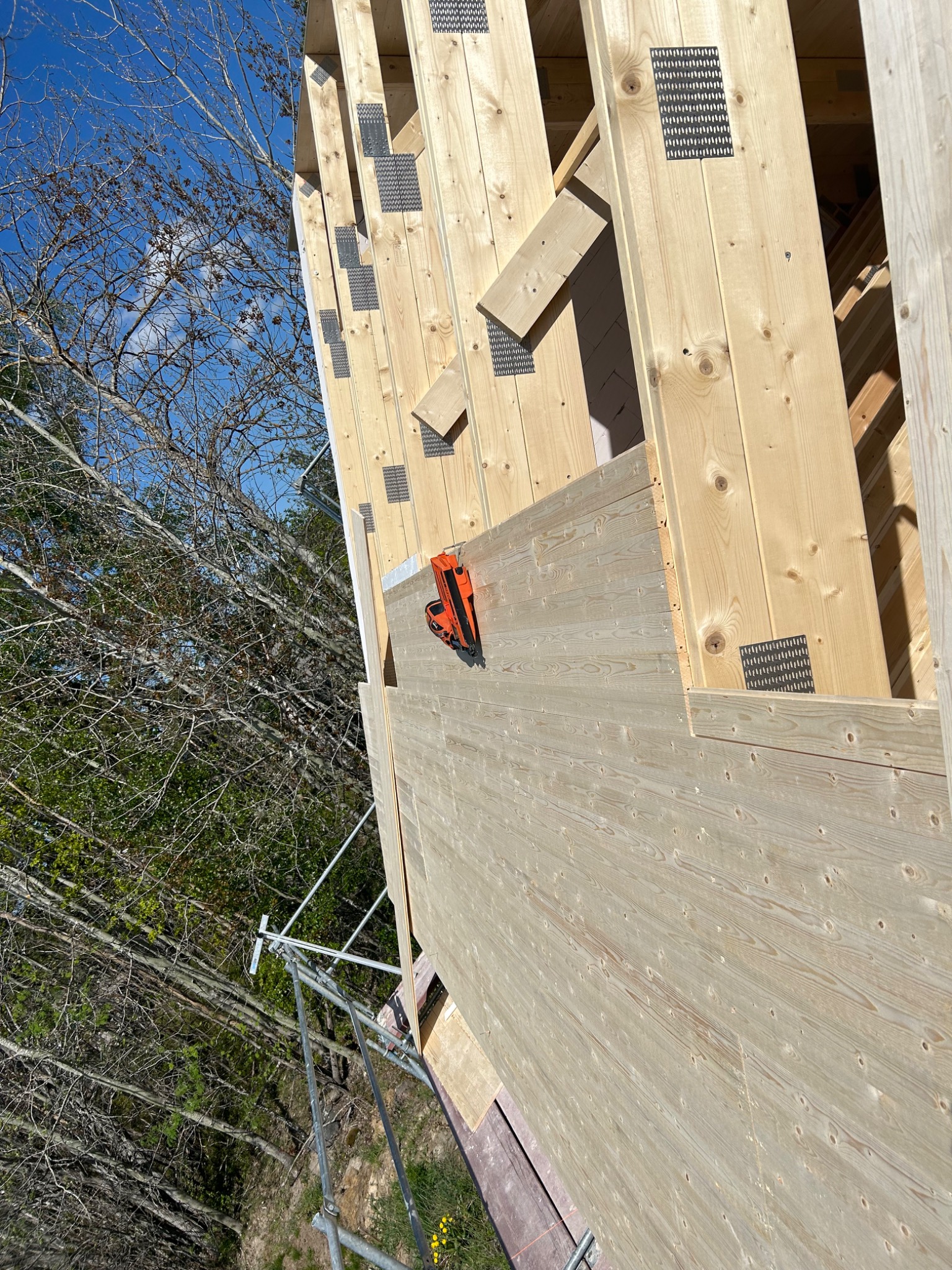 Montaż drewnianej elewacji z widocznym zszywaczem pneumatycznym i elementami konstrukcyjnymi, na tle drzew i błękitnego nieba.