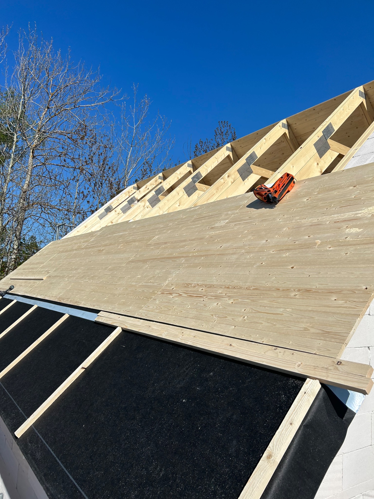 Wysoki kąt widzenia na konstrukcję dachu w trakcie budowy, z widocznymi krokwiami, deskowaniem z desek i membraną dachową, na tle błękitnego nieba i drzew bez liści.