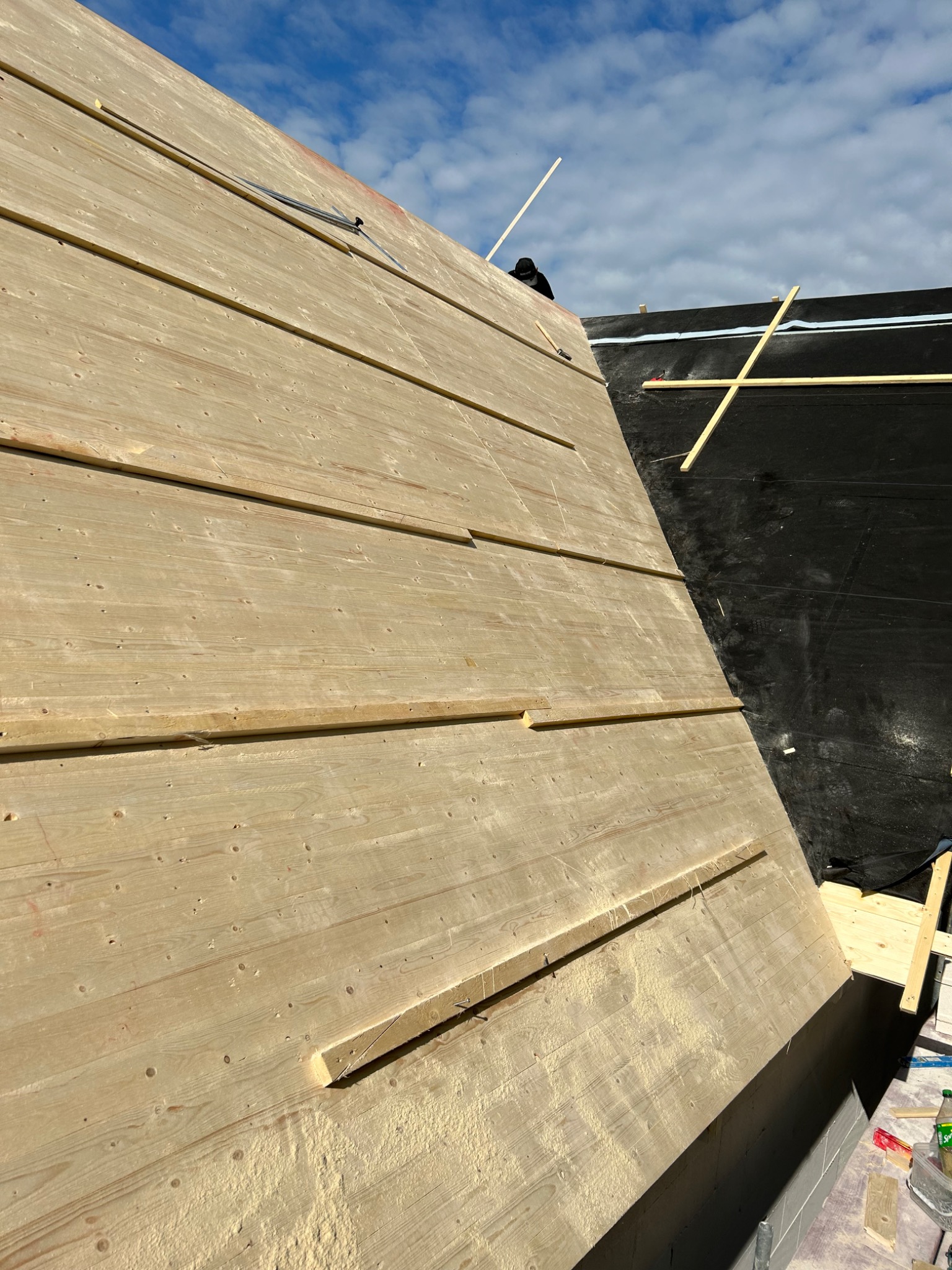 Poddasze w trakcie ocieplania wełną mineralną, widoczne deski mocujące, czarna membrana oraz fragment nieba z chmurami.