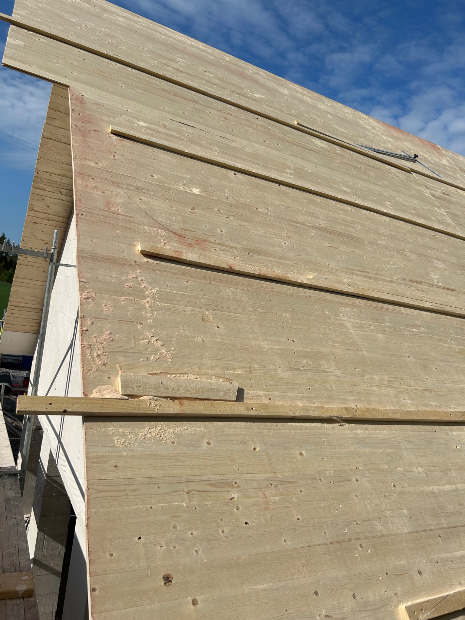 Fragment ściany budynku w trakcie montażu drewnianej elewacji, widoczne deski z jasnego drewna, listwy mocujące i fragment rusztowania.