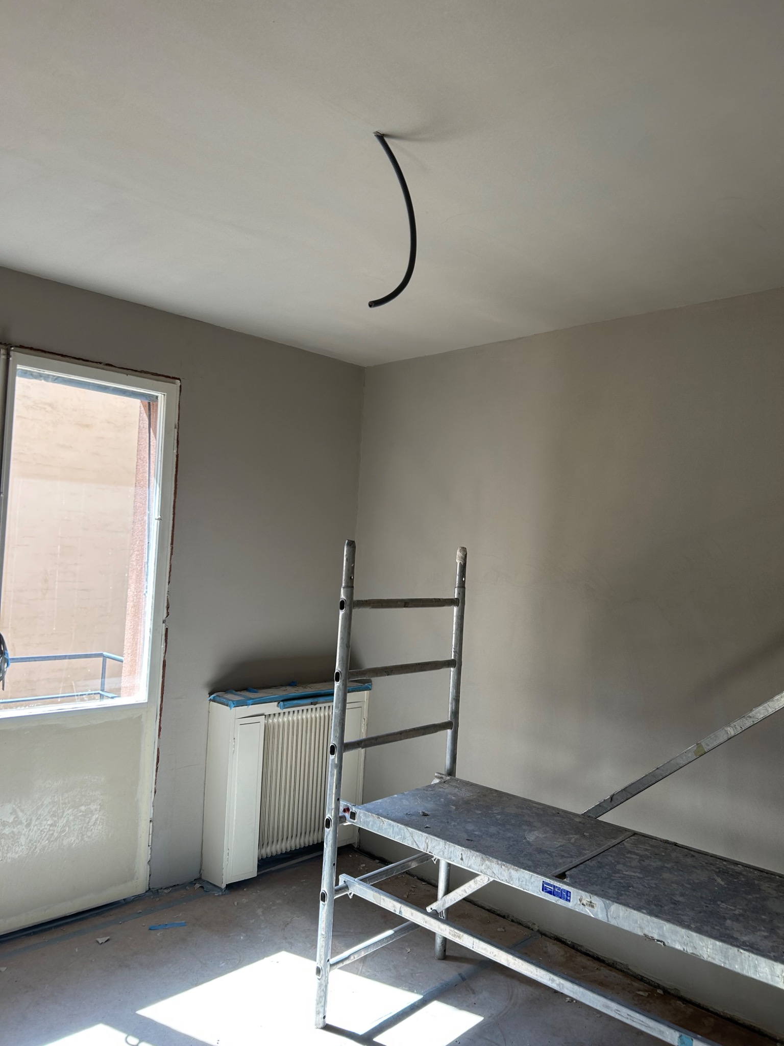 Wnętrze pokoju w trakcie remontu, ściany i sufit świeżo pomalowane na jasny beż, widoczne rusztowanie, wystający kabel z sufitu, okno z widokiem na zewnątrz.