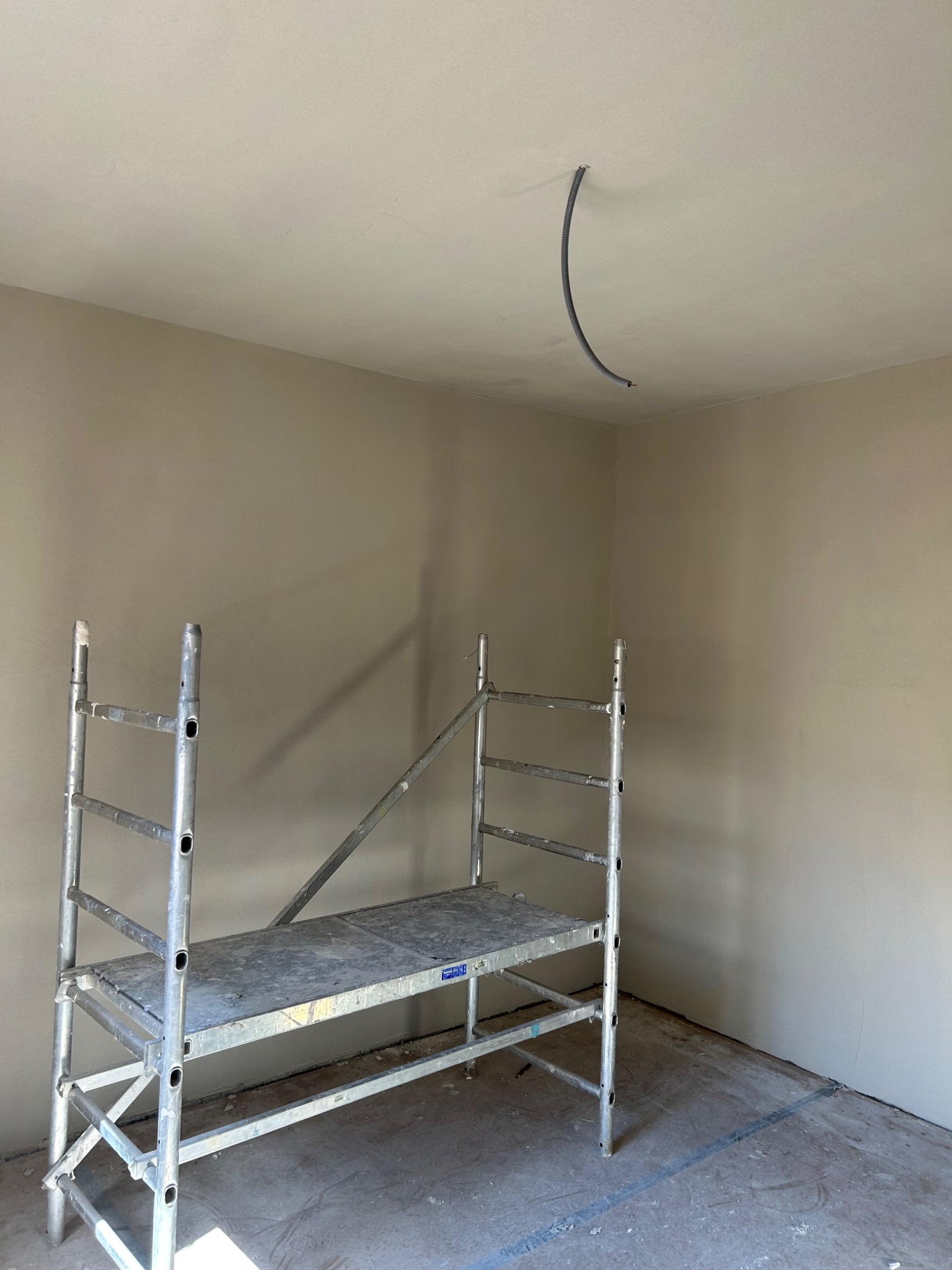 Wnętrze pokoju w trakcie prac wykończeniowych, ściany pokryte świeżym tynkiem w odcieniach beżu, widoczne metalowe rusztowanie i wystający przewód elektryczny z sufitu.