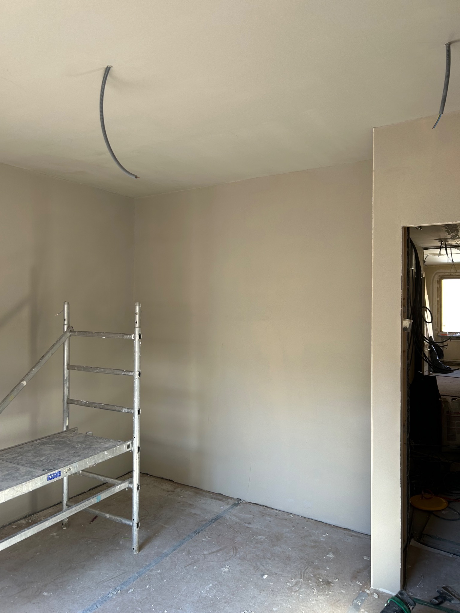 Przygotowane do malowania wnętrze pokoju z metalowym rusztowaniem, wystającymi przewodami elektrycznymi z sufitu i widocznym wejściem do innego pomieszczenia w trakcie remontu.