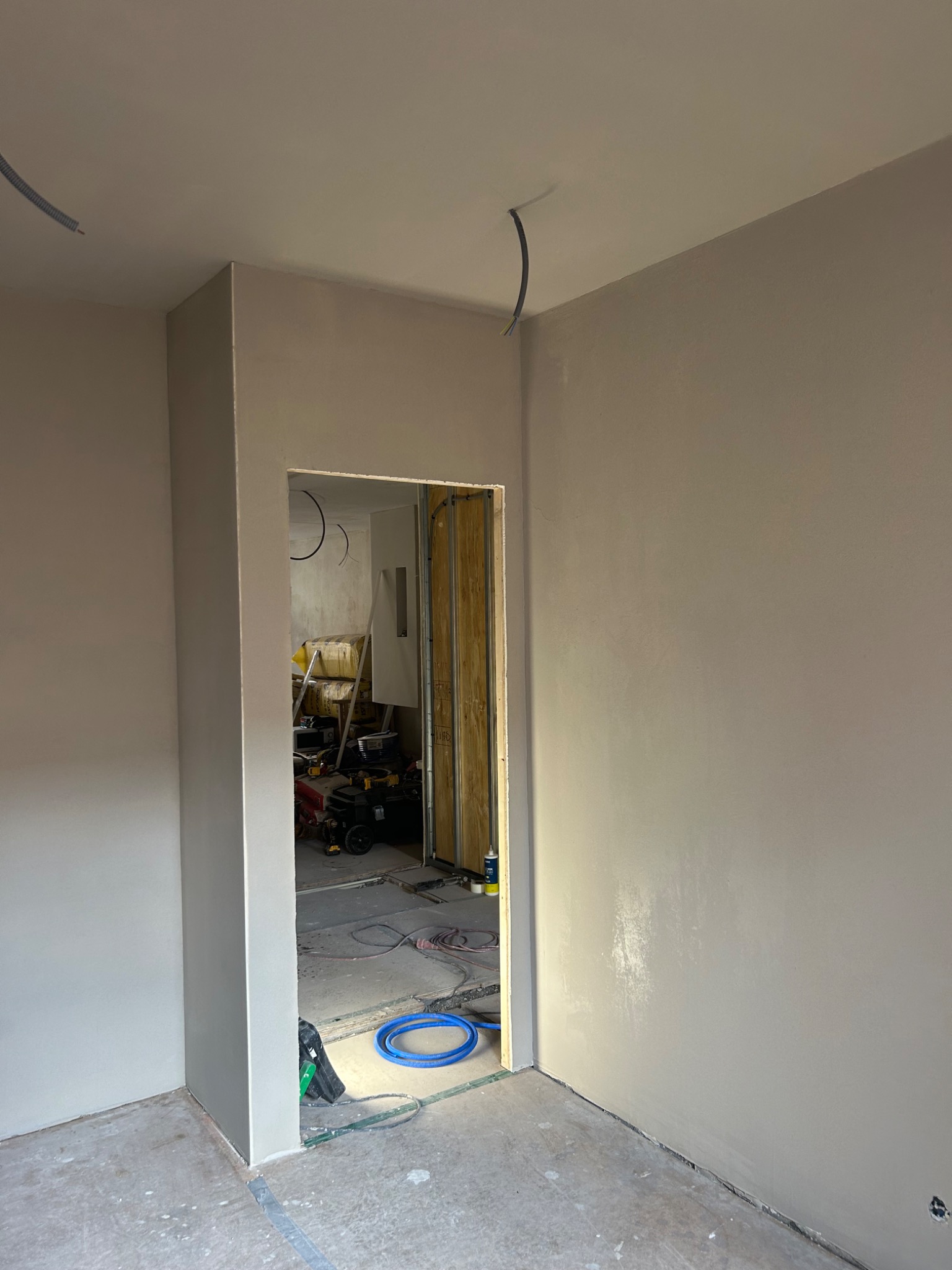 Świeżo otynkowane wnętrze z widocznym otworem drzwiowym, przez który widać narzędzia i materiały budowlane, oraz wystające kable ze ściany.