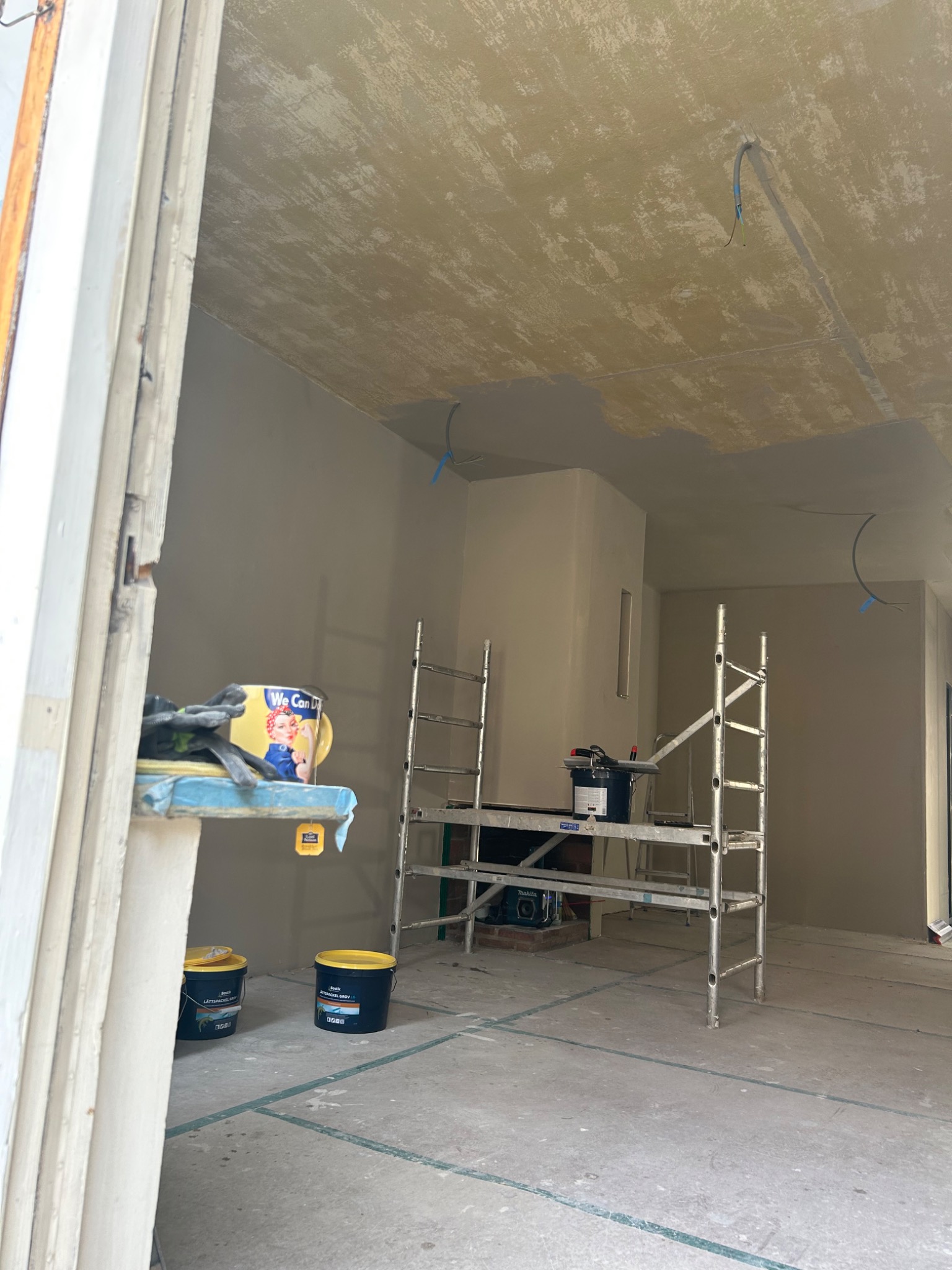 Wnętrze w trakcie malowania: sufit z widocznymi śladami szpachlowania, ściany pomalowane na jasny kolor, rozstawiony metalowy ruszt, puszki z farbą na podłodze i charakterystyczna puszka z motywem...