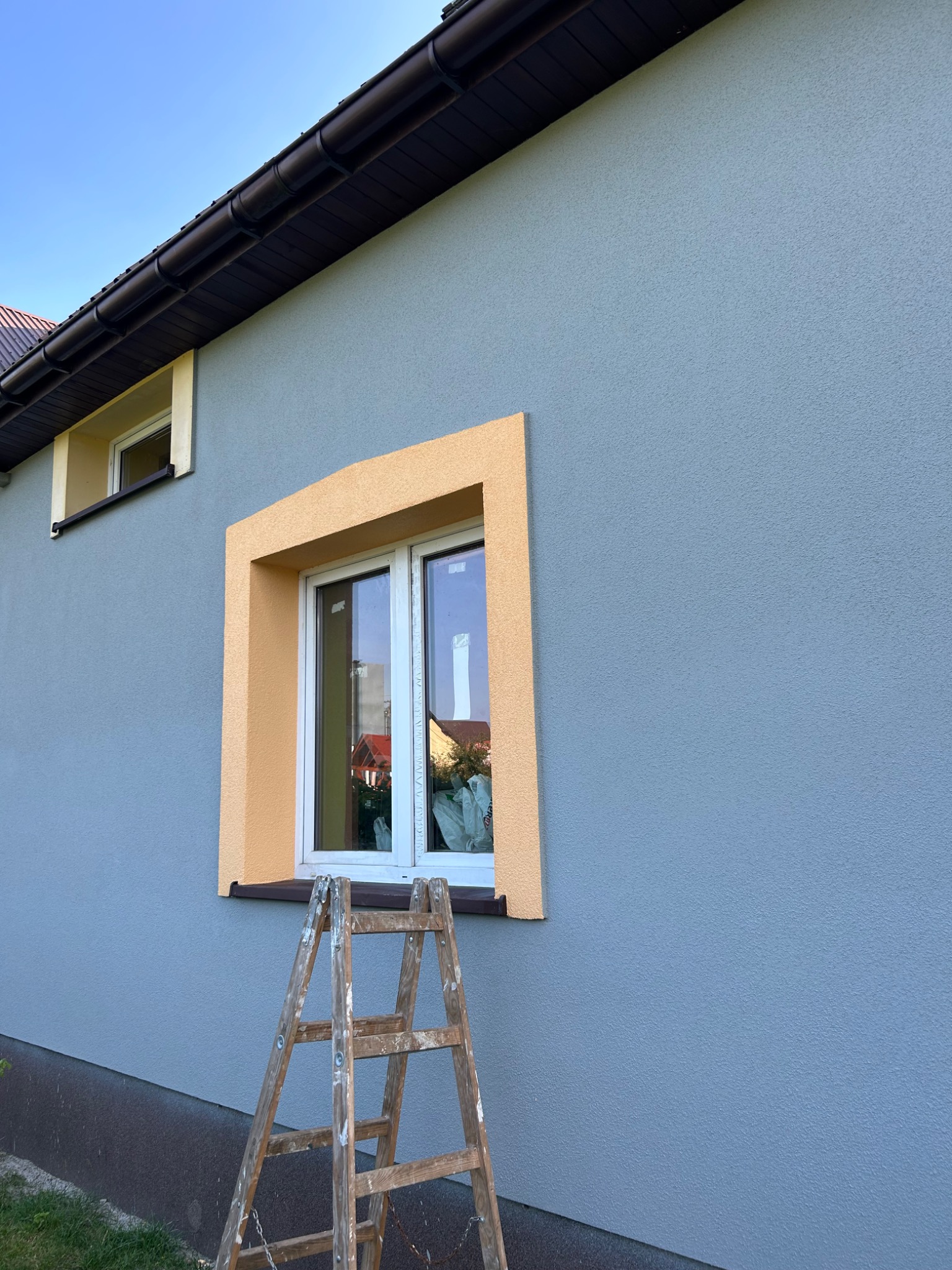 Ściana budynku z oknem w żółtej ramie, widoczna drewniana drabina, świeżo położony tynk w odcieniach szarości i żółci.
