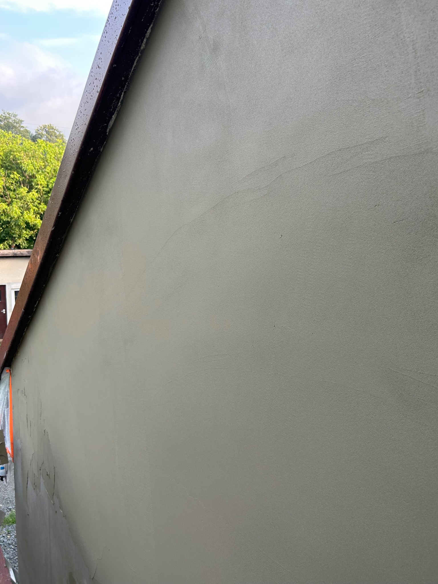 Świeżo otynkowana ściana budynku z widocznym fragmentem dachu, wykonana w jasnym odcieniu szarości, z widocznymi śladami po narzędziach i lekkimi smugami na powierzchni.