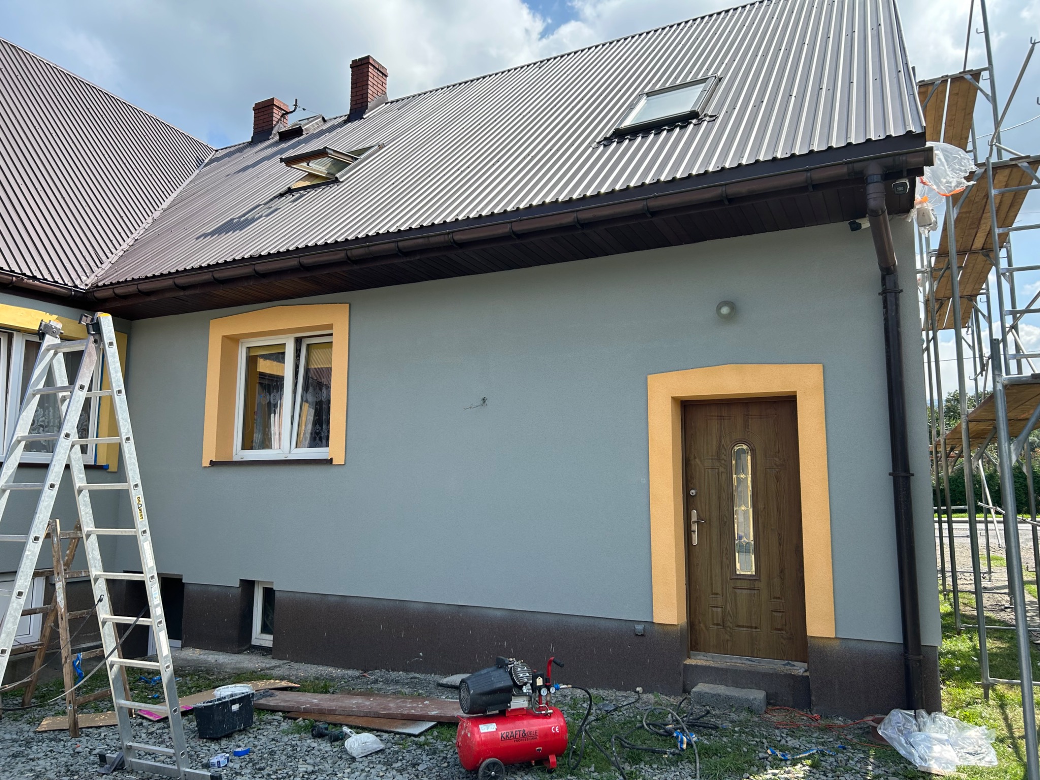Szary dom jednorodzinny z nową elewacją, brązowym dachem z oknami połaciowymi i stolarką okienną oraz drzwiową w kolorze żółtym, widoczny kompresor i drabina