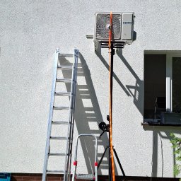 JM KLIMATYZACJA - Montaż klimatyzatora Heiko Brisa na elewacji budynku. Widoczne drabiny, podnośnik i narzędzia. Słoneczny dzień, trawa i fragment okna w tle.