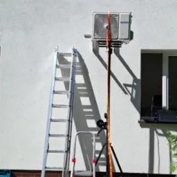 Montaż klimatyzatora Heiko Brisa na elewacji budynku. Widoczne drabiny, podnośnik i narzędzia. Słoneczny dzień, trawa i fragment okna w tle.