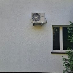 JM KLIMATYZACJA - Biała jednostka zewnętrzna klimatyzacji Heiko, zamontowana na ścianie budynku z oknem, widok z poziomu trawnika.
