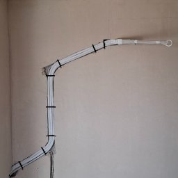 JM KLIMATYZACJA - Białe rury instalacji, przymocowane czarnymi opaskami do ściany, połączone z gniazdkiem elektrycznym. Na podłodze worek i odkurzacz.