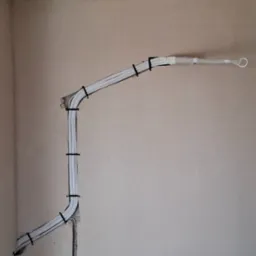 Białe rury instalacji, przymocowane czarnymi opaskami do ściany, połączone z gniazdkiem elektrycznym. Na podłodze worek i odkurzacz.