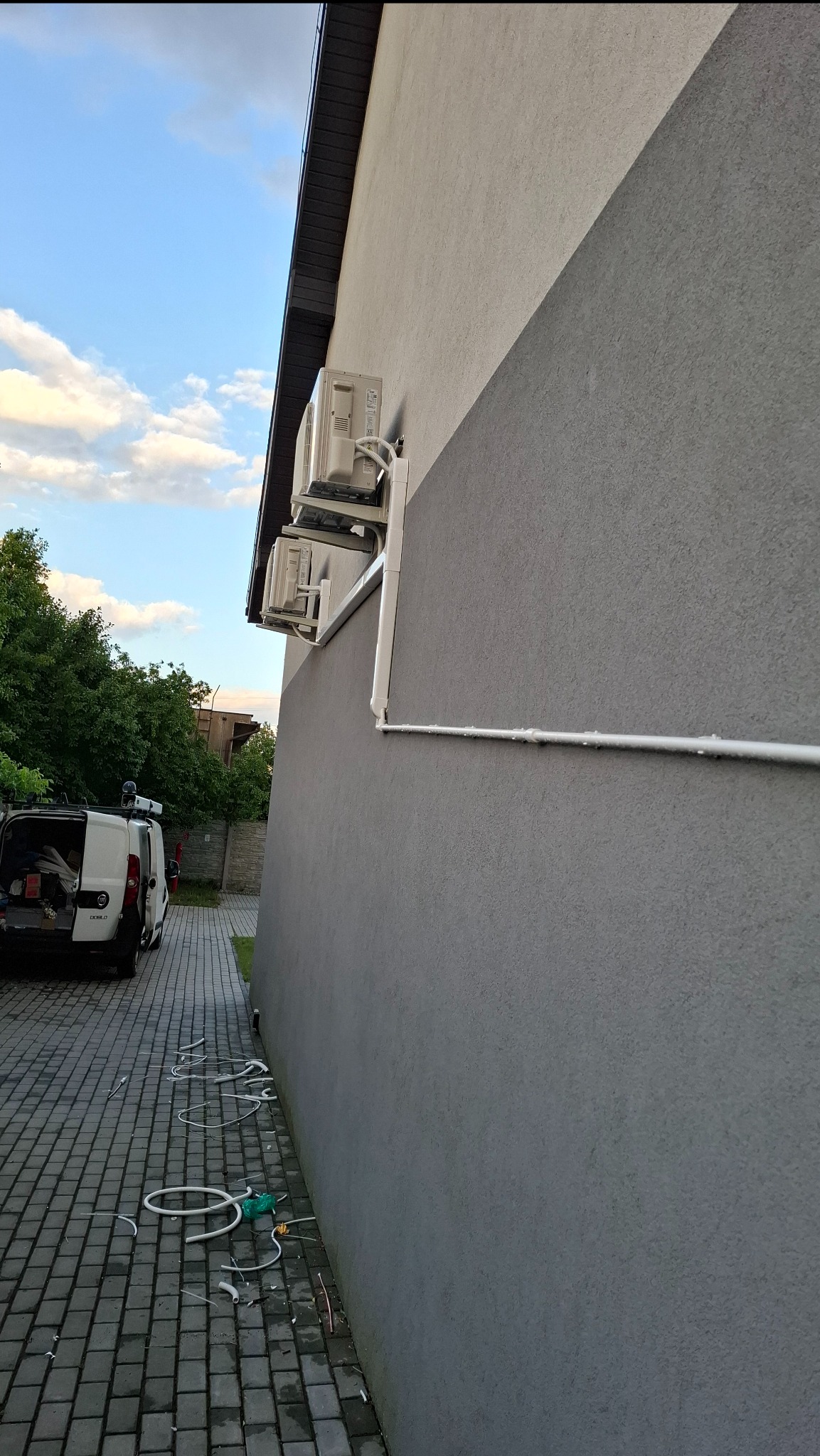 Dwa klimatyzatory na szarej ścianie budynku, białe rury odprowadzające skropliny, samochód dostawczy w tle, kostka brukowa i fragmenty kabli na ziemi.