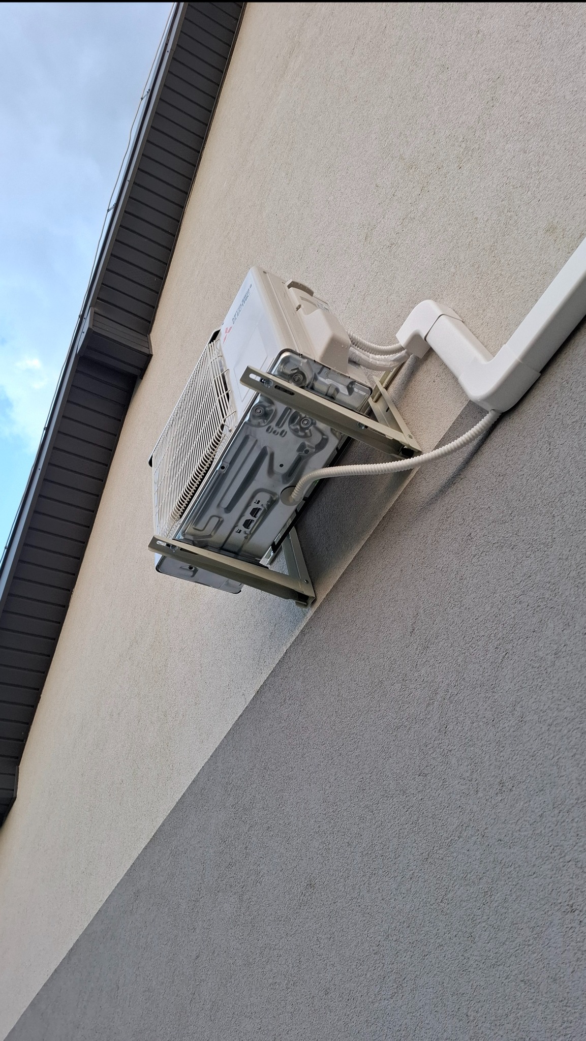 Biały klimatyzator Mitsubishi zamontowany na elewacji budynku, widoczny z dołu, z przewodami i rurami odprowadzającymi. Częściowo widoczne orynnowanie.