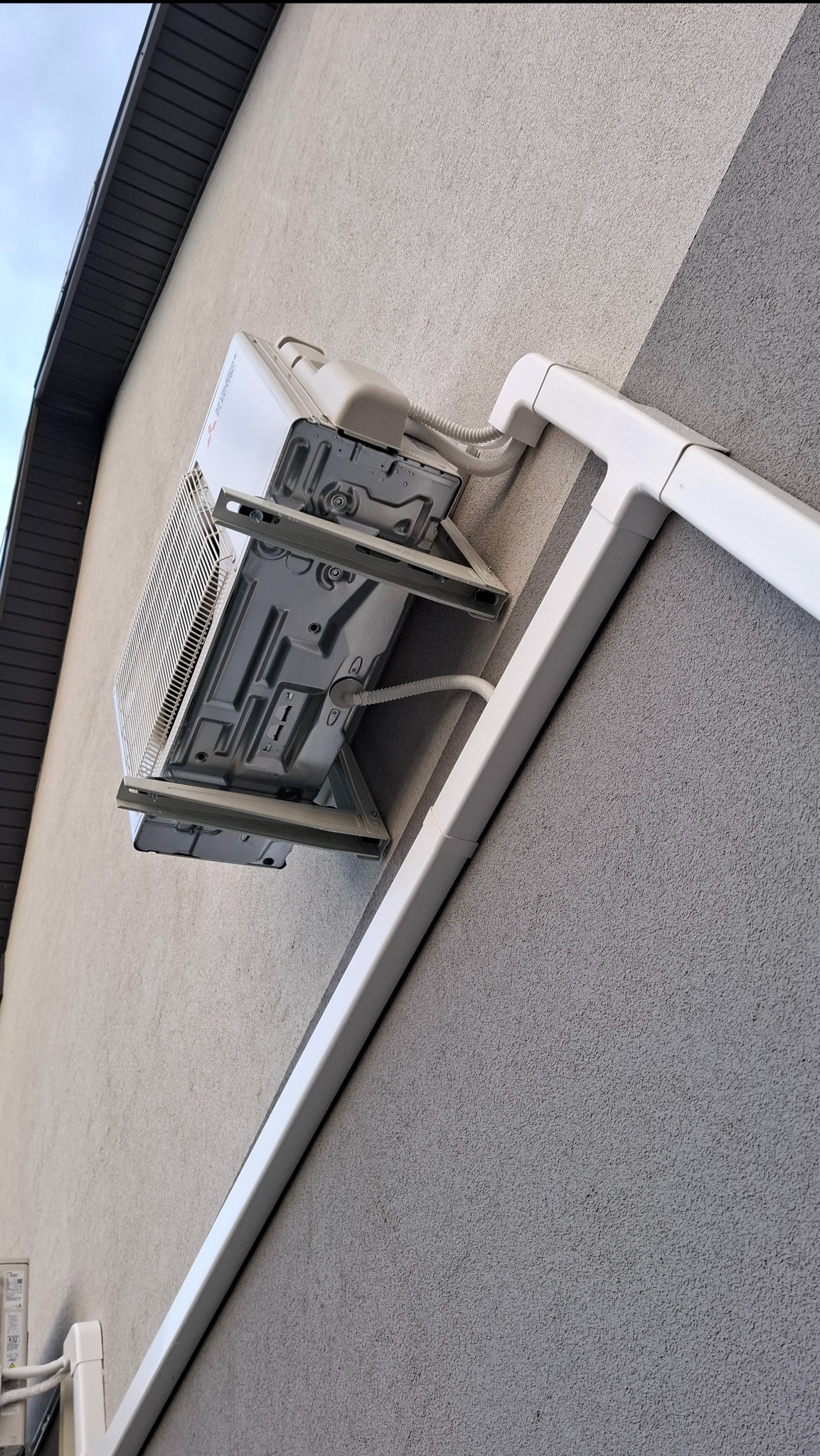 Biała jednostka zewnętrzna klimatyzacji Mitsubishi zamontowana na szarej, teksturowanej ścianie budynku z widocznymi białymi kanałami instalacyjnymi.