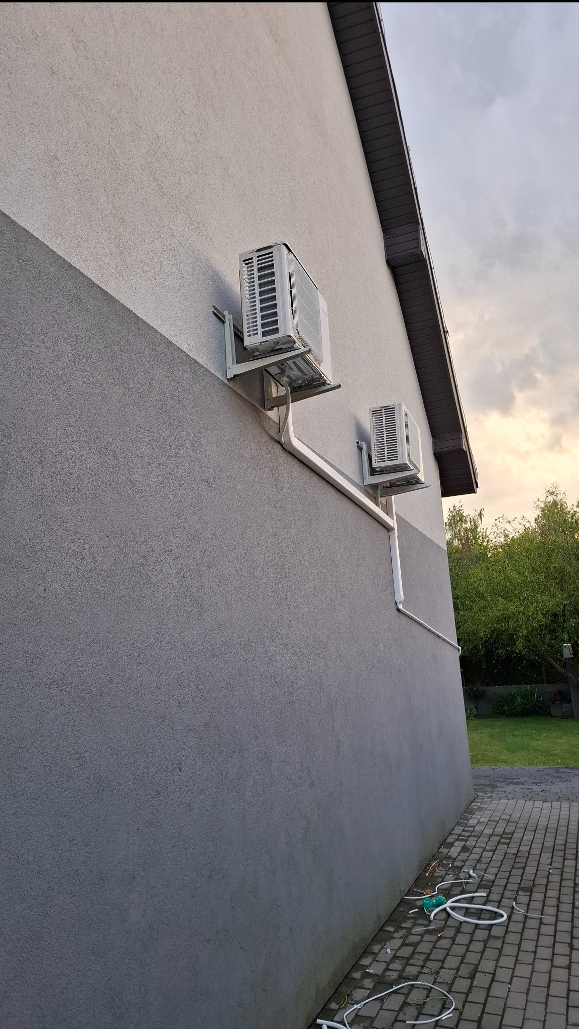 Dwa białe klimatyzatory zamontowane na szarej ścianie budynku z widoczną instalacją odprowadzającą skropliny. Widok z dołu, z boku budynku.