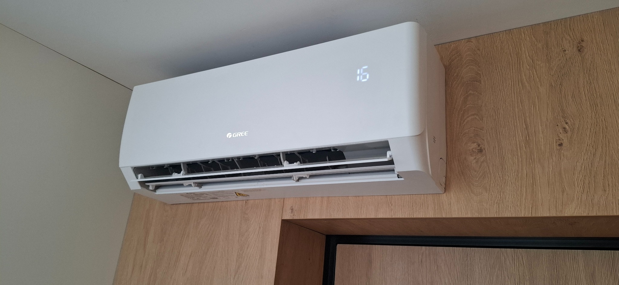 Biała jednostka klimatyzacji Gree Pular zamontowana na ścianie w pomieszczeniu. Widoczny wyświetlacz temperatury i logo producenta.