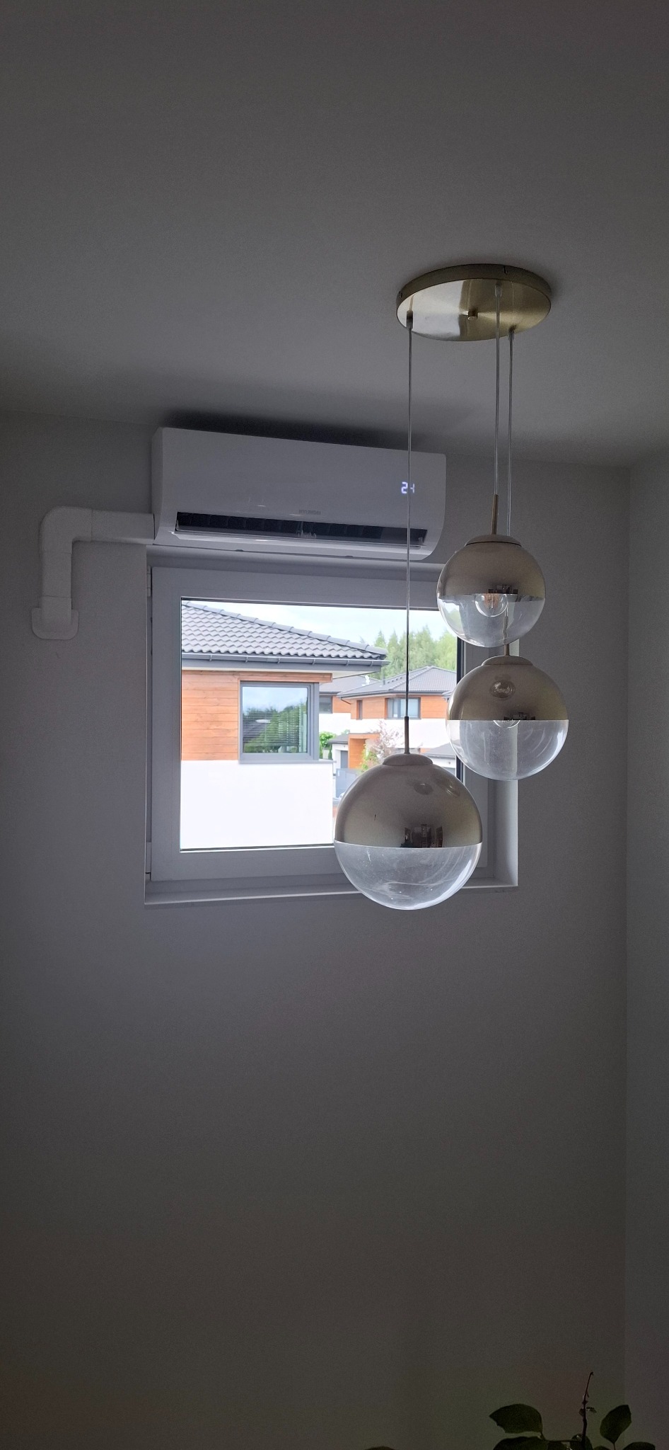 Biały klimatyzator Hyundai Revolution multi zamontowany nad oknem w jasnym wnętrzu, widoczne rury instalacyjne i nowoczesna lampa wisząca.