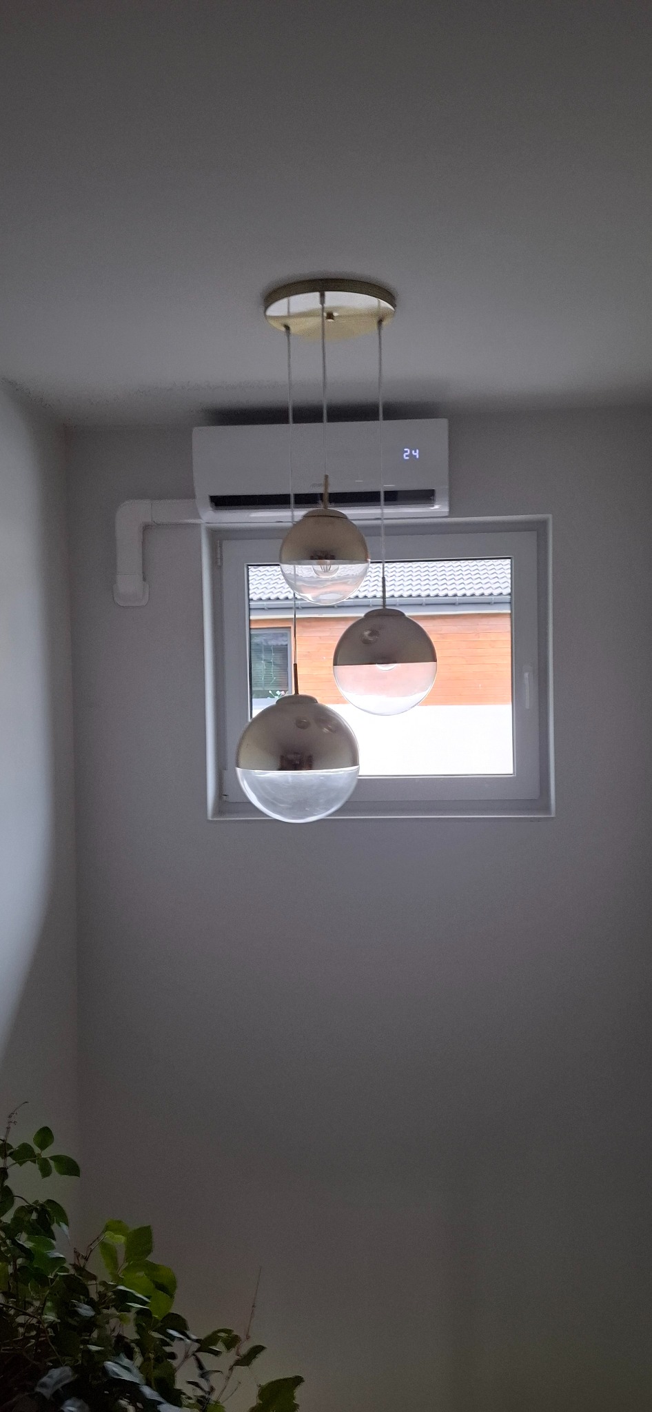 Biała klimatyzacja ścienna nad oknem, widoczna temperatura 24 stopnie, w tle żyrandol z trzema szklanymi kloszami w odcieniach złota. Minimalistyczny design wnętrza.