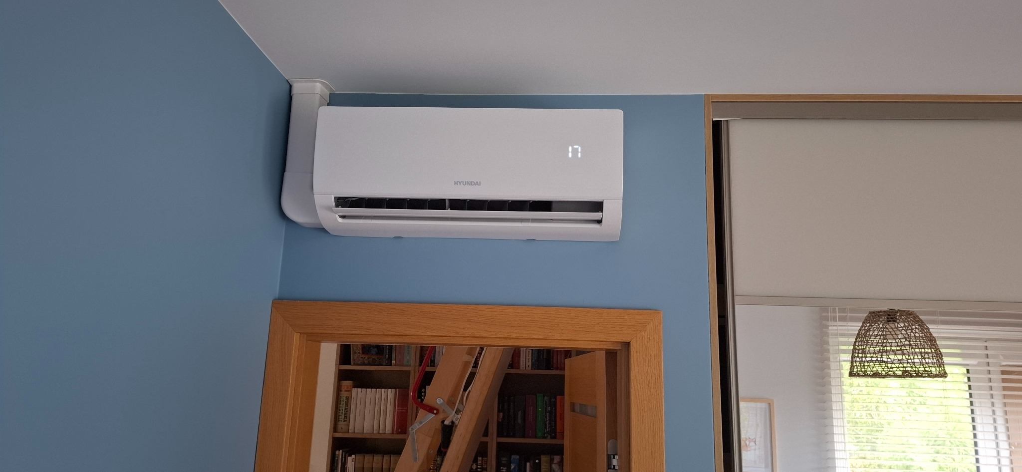 Biały klimatyzator Hyundai na błękitnej ścianie, z widocznym wyświetlaczem temperatury. W tle drzwi z regałem pełnym książek i fragment okna z roletami.