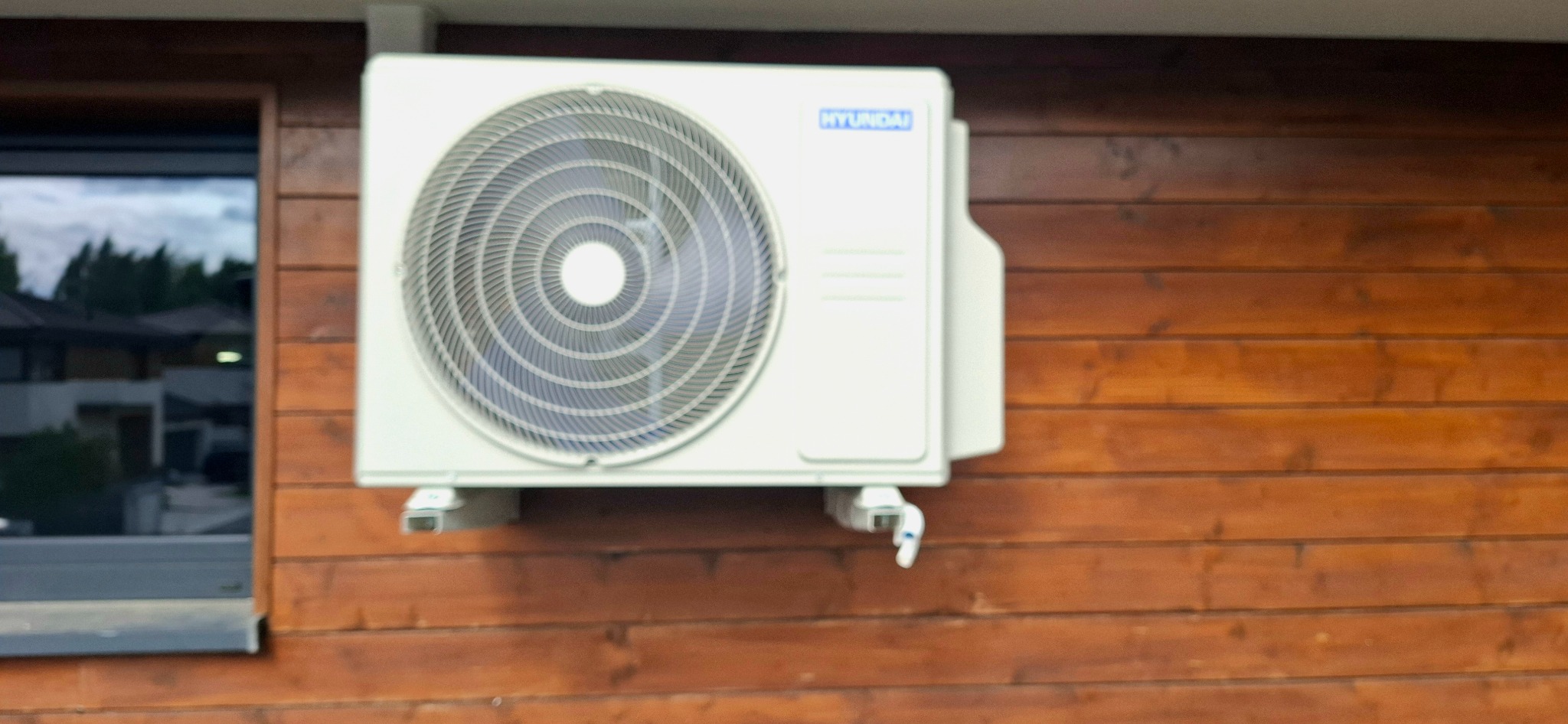 Biała jednostka zewnętrzna klimatyzacji Hyundai zamontowana na brązowej, drewnianej elewacji domu. Widoczny fragment okna z odbijającym się otoczeniem.