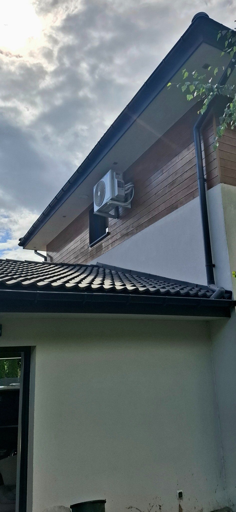 Biała jednostka klimatyzacji zamontowana na drewnianej ścianie domu, widoczna z dołu pod kątem, z fragmentem dachu i błękitnym niebem w tle.