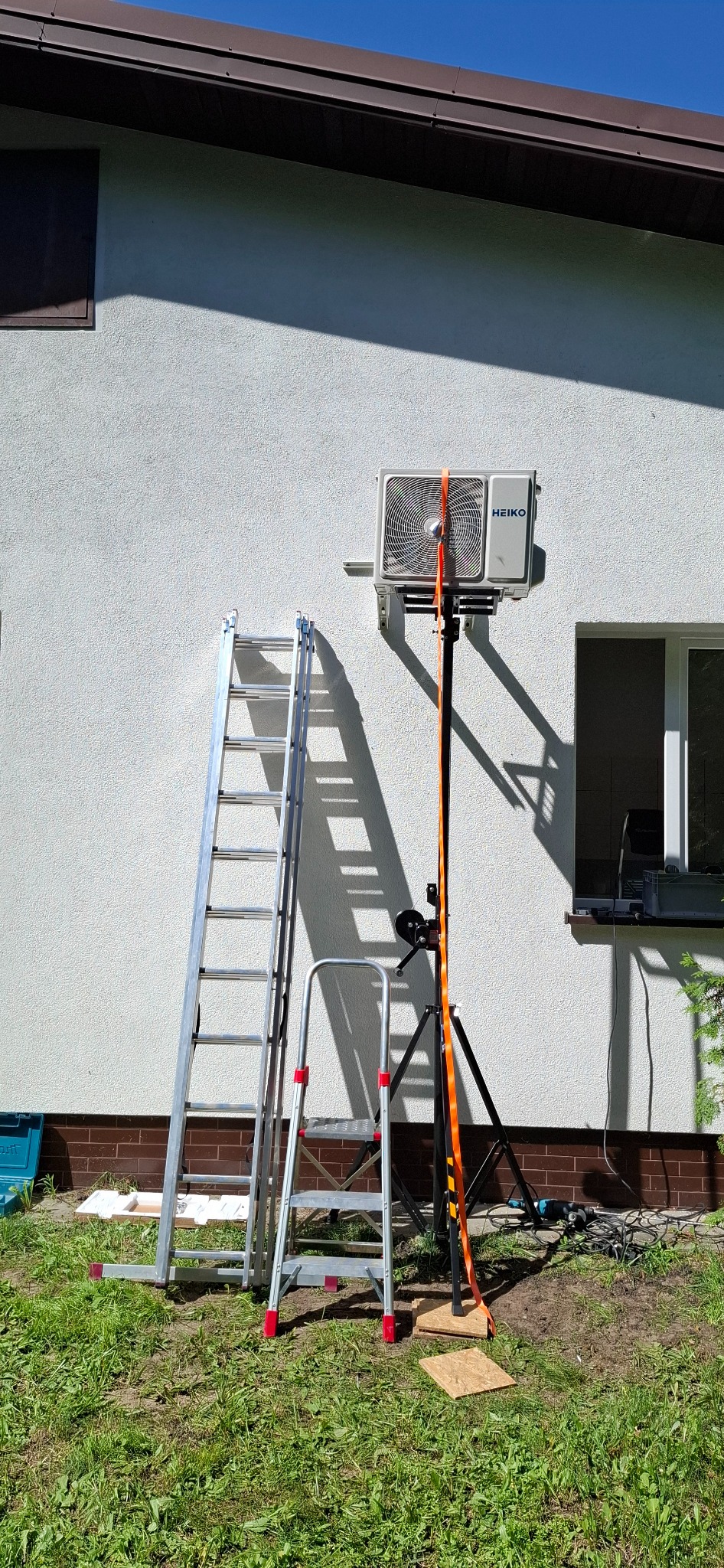 Montaż klimatyzatora Heiko Brisa na elewacji budynku. Widoczne drabiny, podnośnik i narzędzia. Słoneczny dzień, trawa i fragment okna w tle.