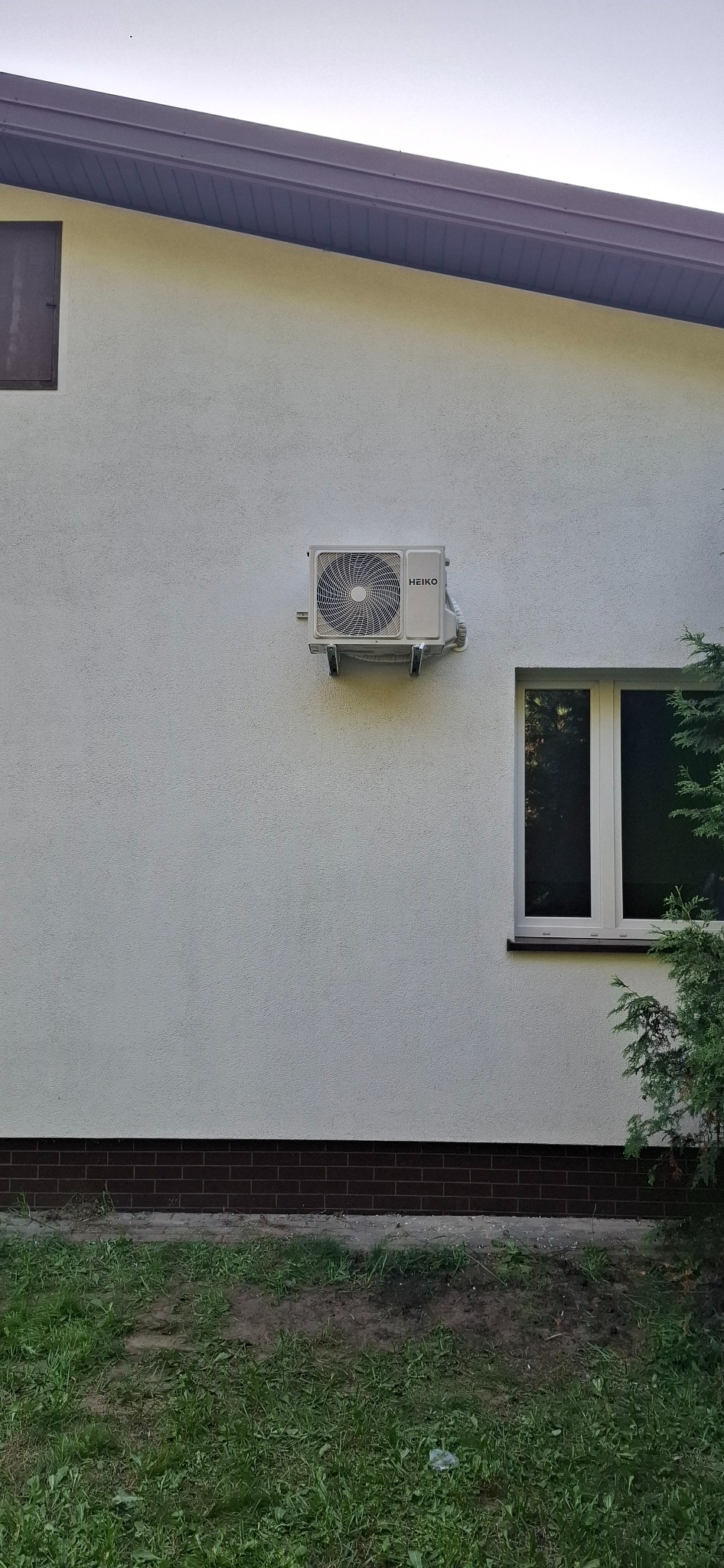 Biała jednostka zewnętrzna klimatyzacji Heiko, zamontowana na ścianie budynku z oknem, widok z poziomu trawnika.